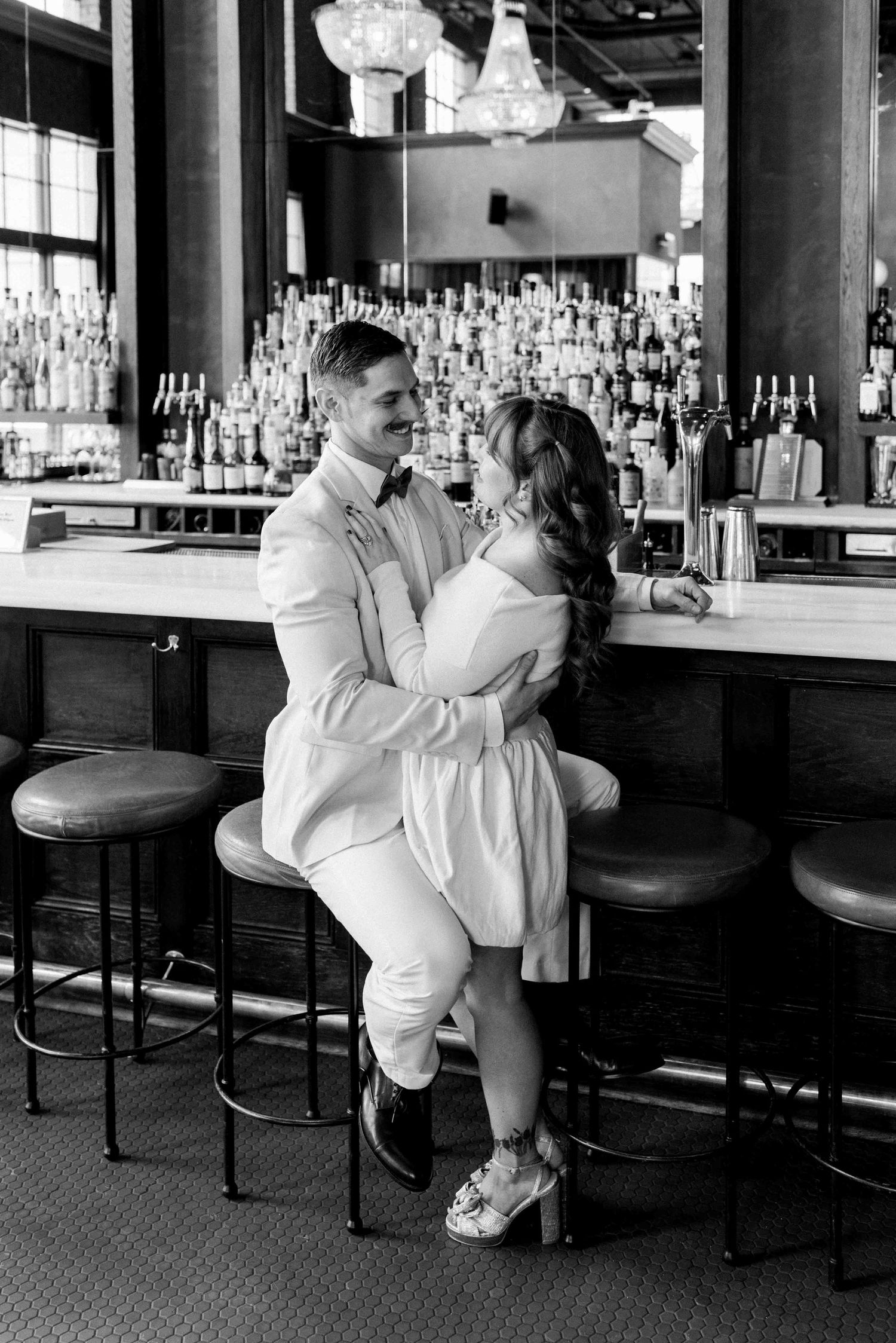 A romantic couple embraces at an upscale bar with rows of liquor bottles in the background.