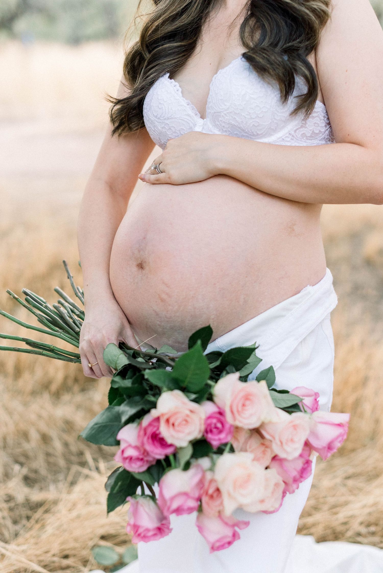 Close-up maternity photo featuring pink rose bouquet and white lace in golden light.