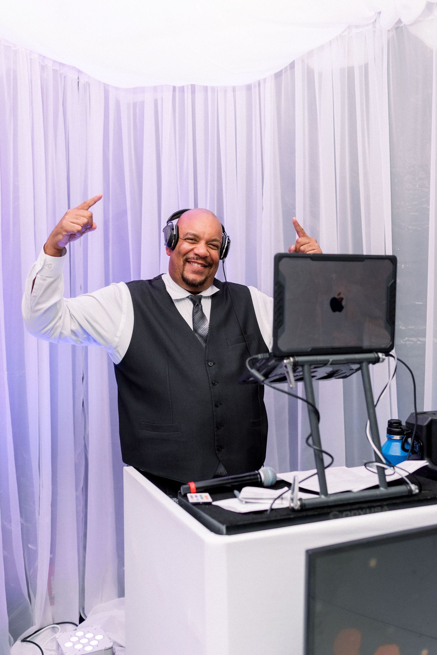 DJ performs enthusiastically with laptop at wedding reception against white backdrop.