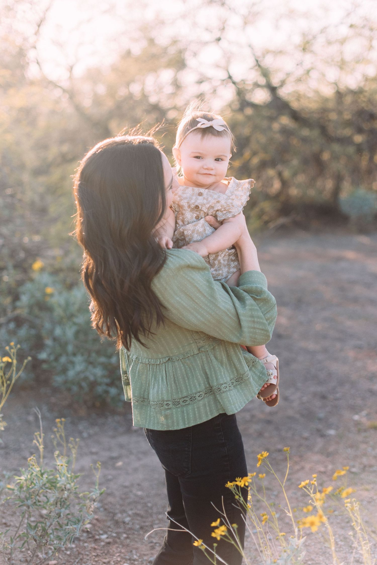 Mother in green dress holds child while standing on desert trail with yellow flowers.