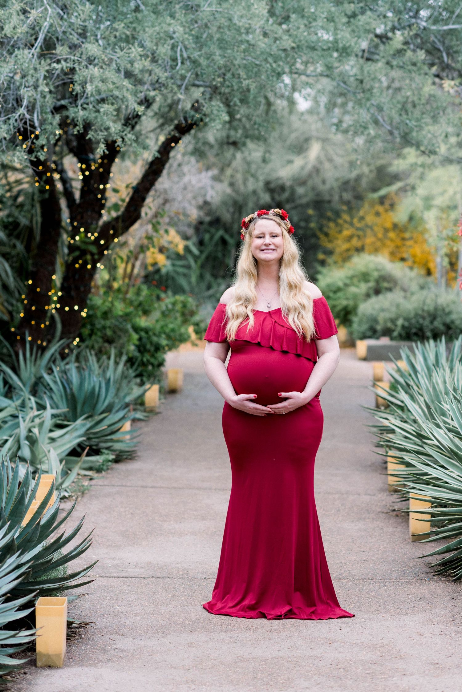 Maternity portrait in flowing red gown on garden path with agave plants and luminarias.