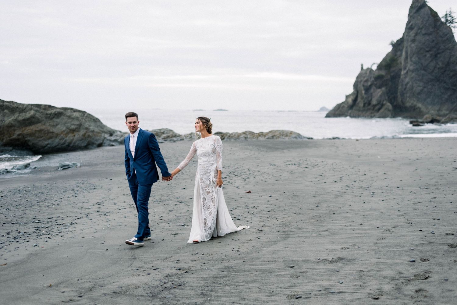 A couple in wedding attire walks along a moody Pacific Northwest beach with rocky cliffs.