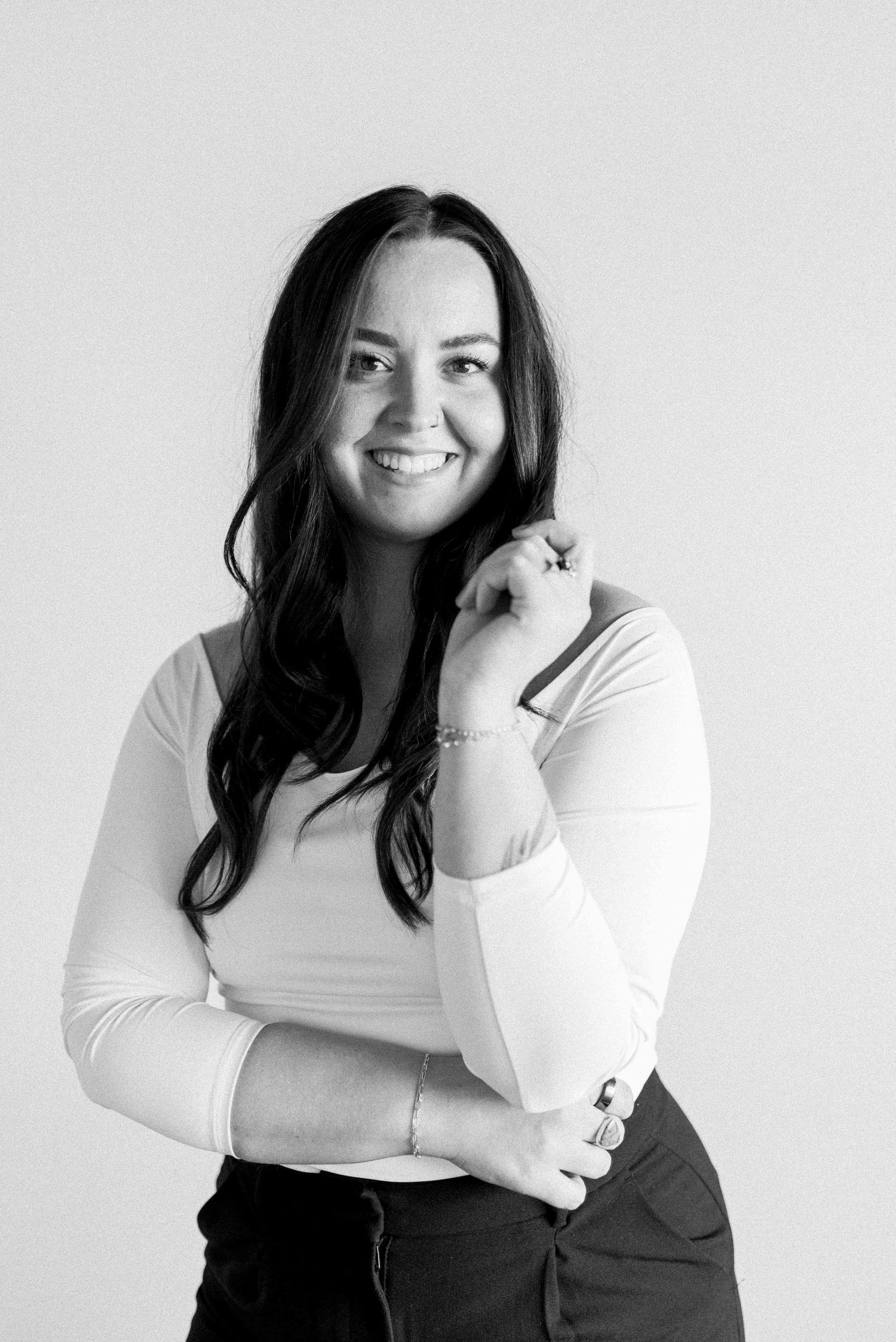 Black and white portrait of a woman with long dark hair wearing a white long sleeve top and dark pants against light background.