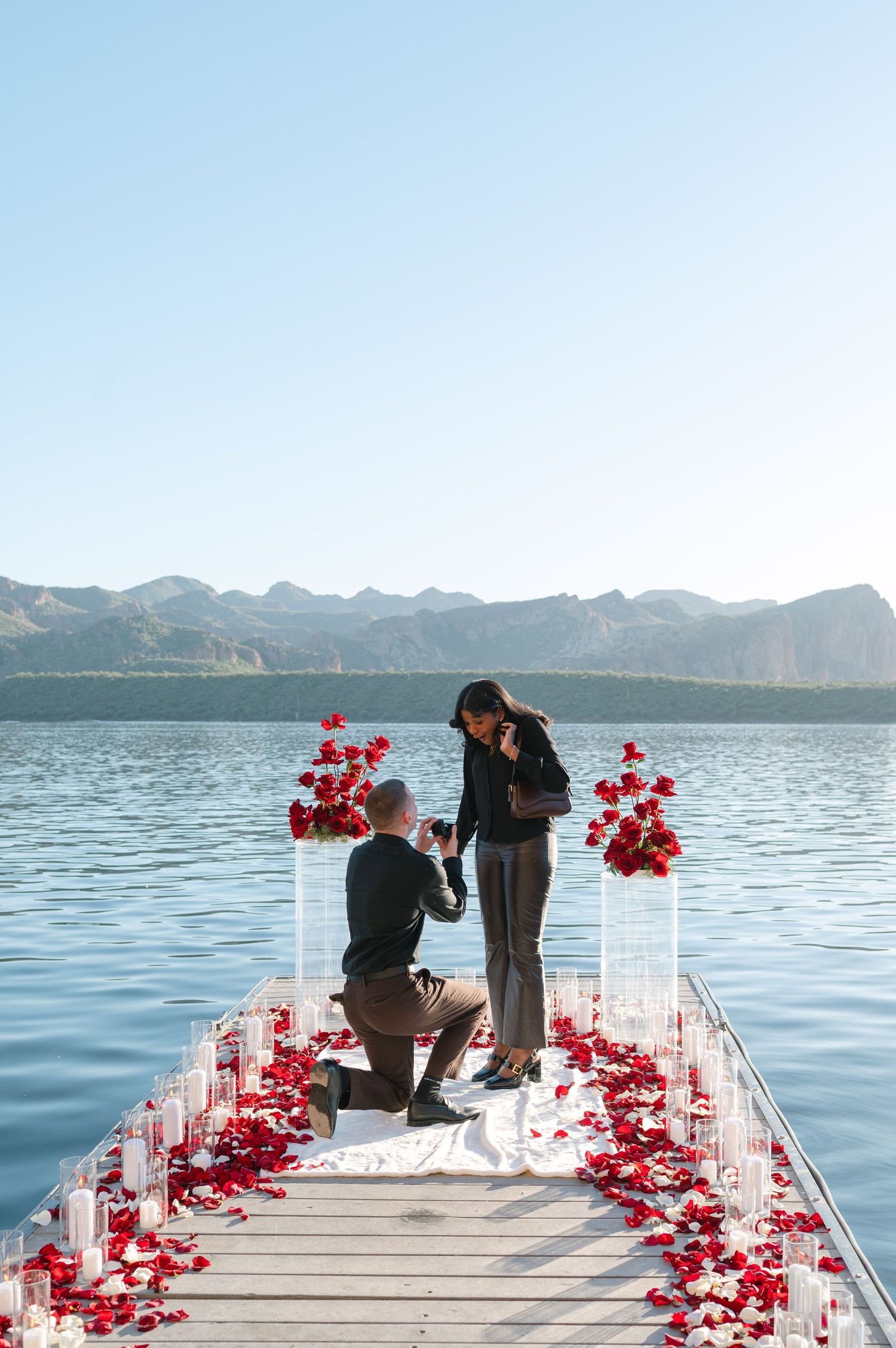 A romantic proposal setup on a lakeside dock with red rose petals and white pillars against a mountain backdrop.