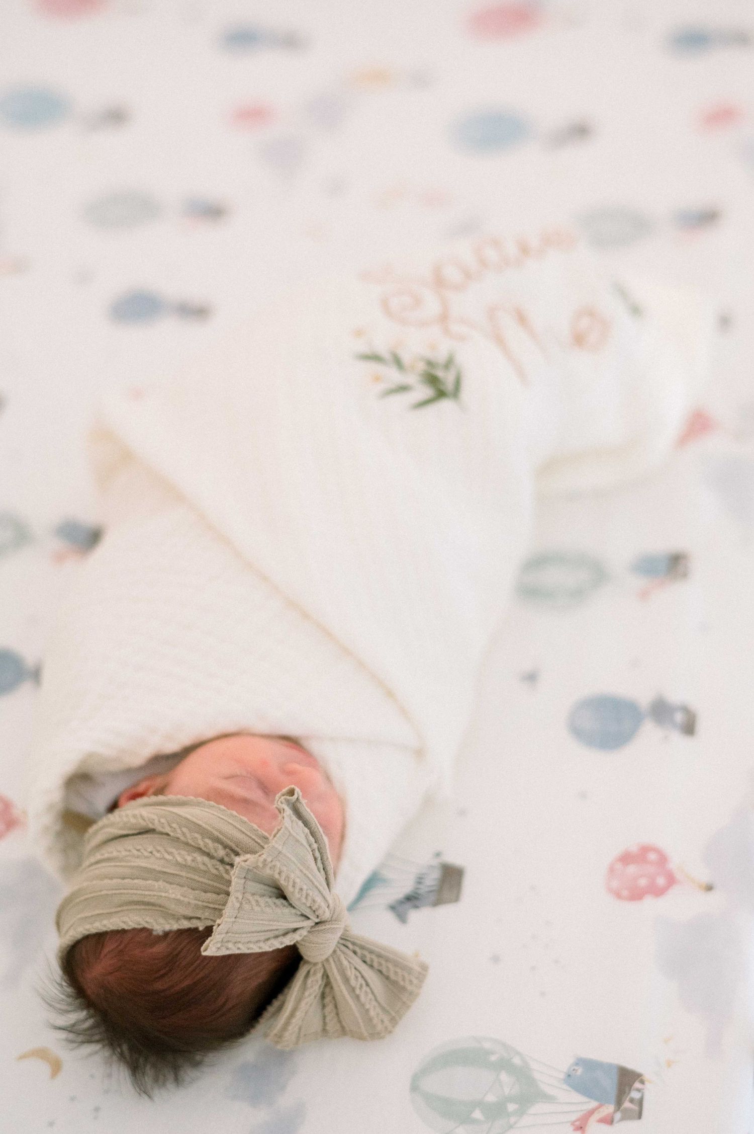 Newborn baby wearing a beige bow headband rests on patterned bedding.