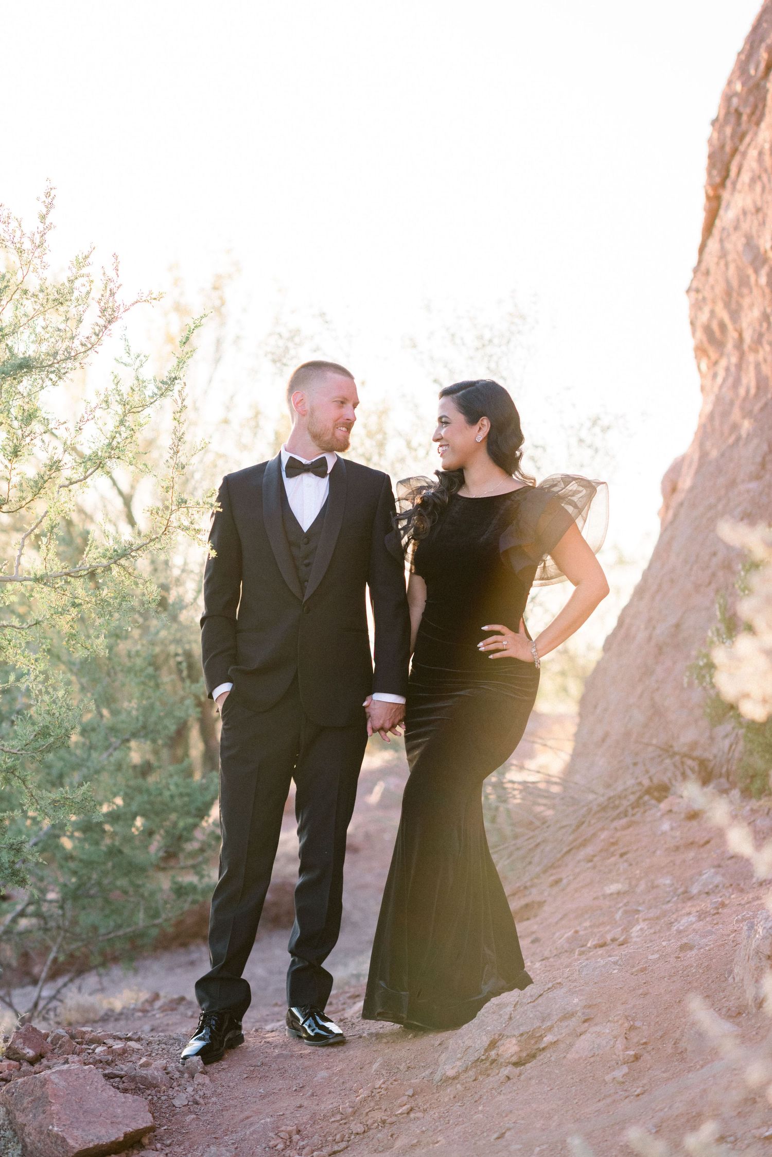Stylish couple in black formal wear standing against rocky desert landscape.