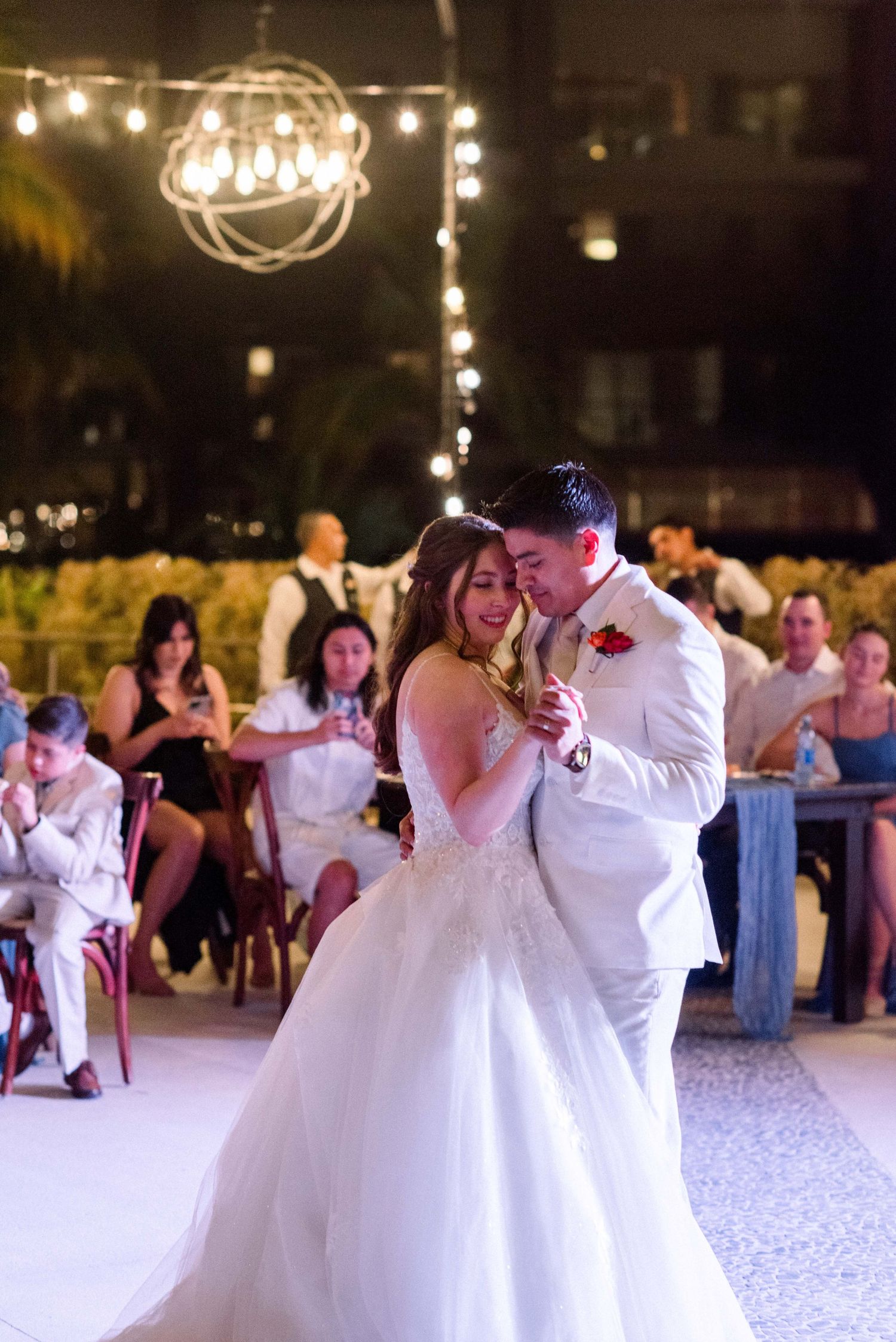 Wedding couple slow dancing under glowing string lights at evening reception.