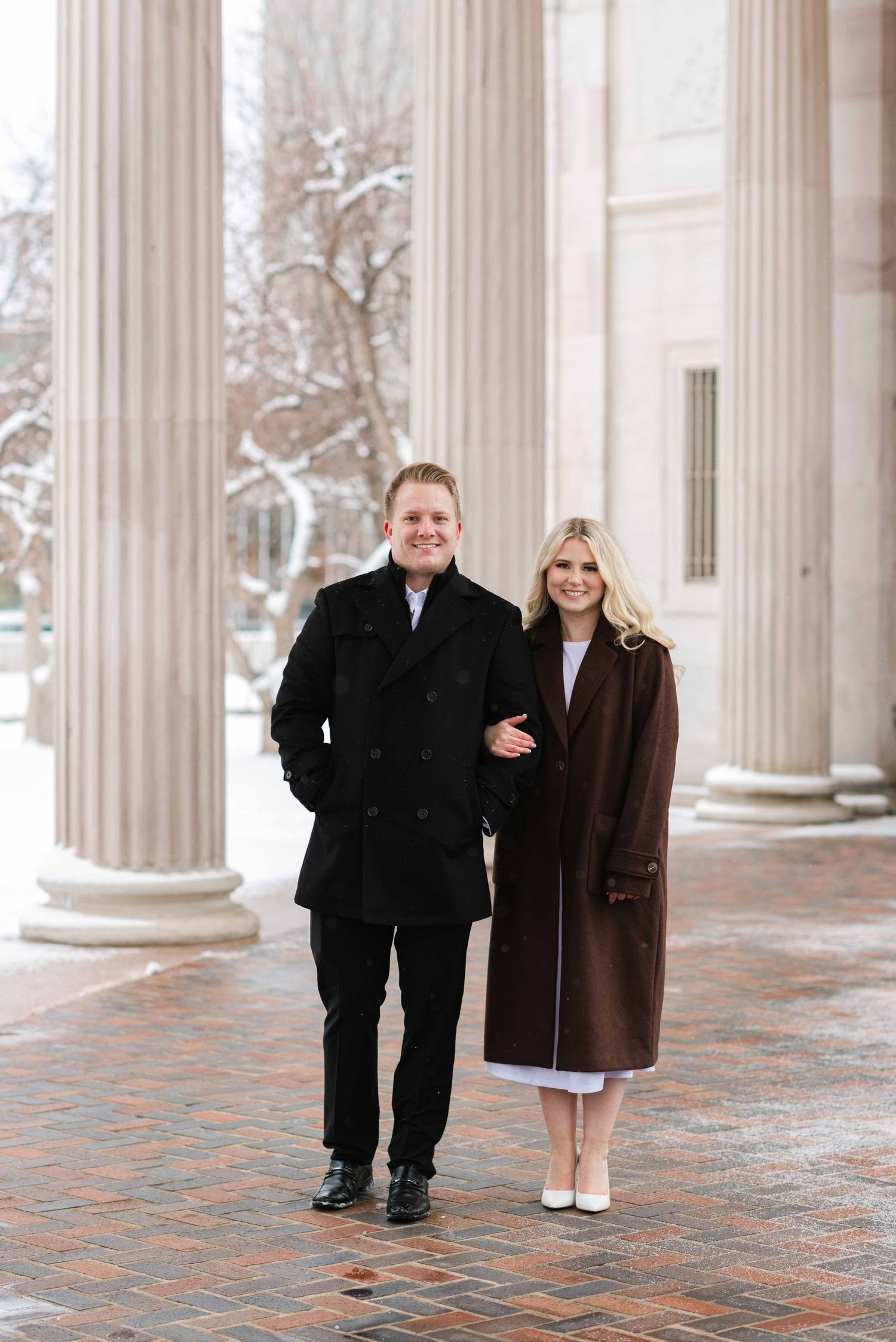 A couple in winter coats poses between classical columns on a snowy day.