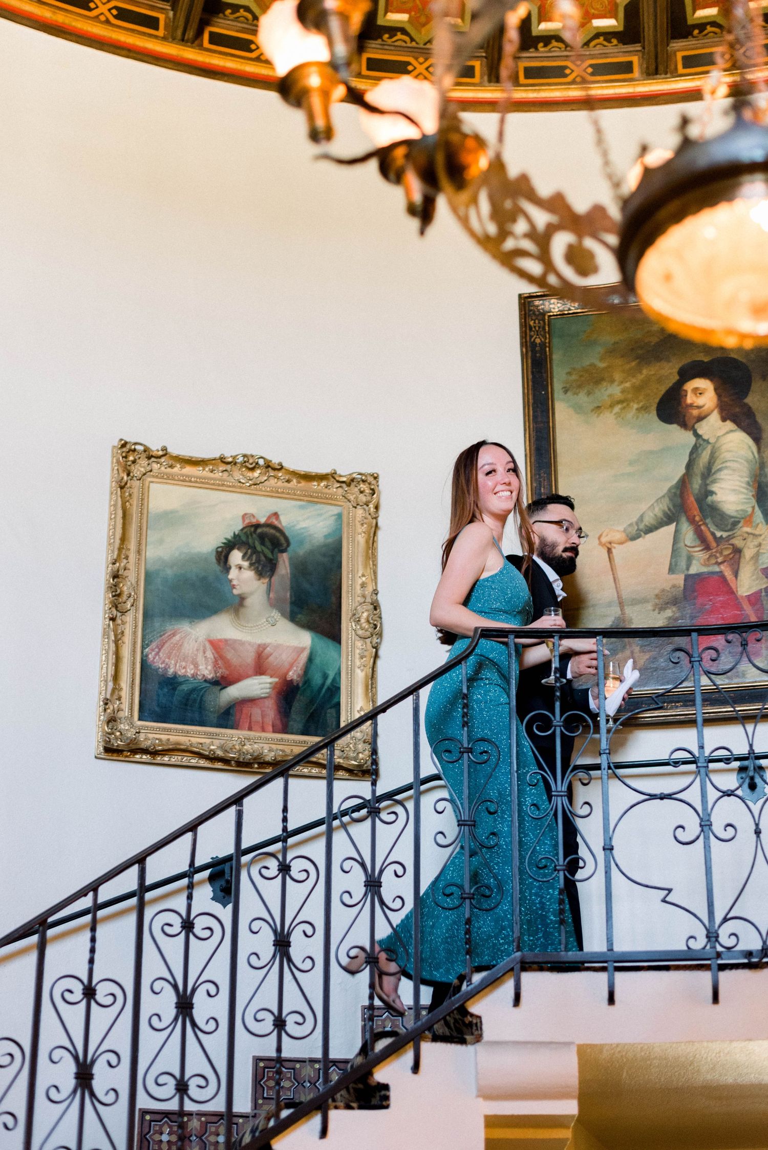 Couple poses on elegant staircase with ornate frames and chandelier in background.
