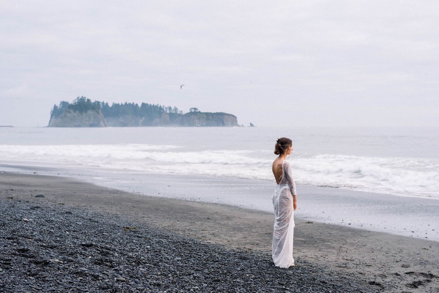 Person in white dress stands alone on a black sand beach looking at distant sea cliffs.