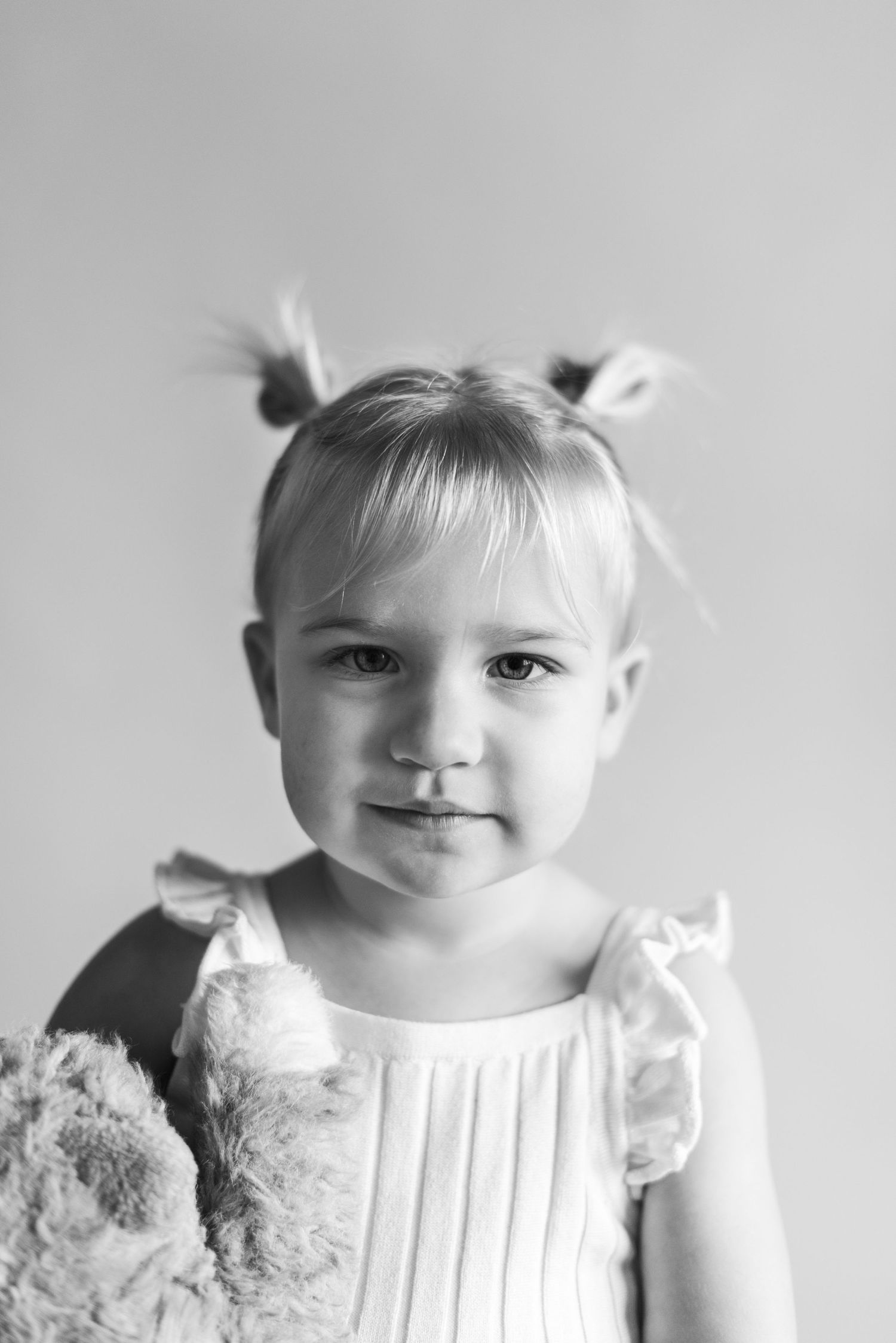 Portrait of a small child in white dress with hair in buns against light background.