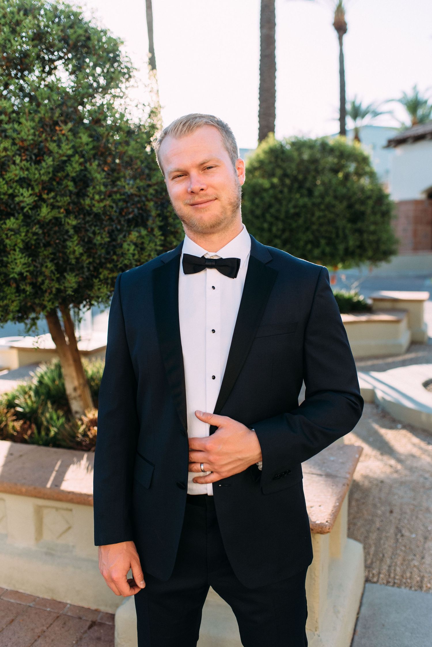 A groom in a black tuxedo poses outdoors against a garden background.