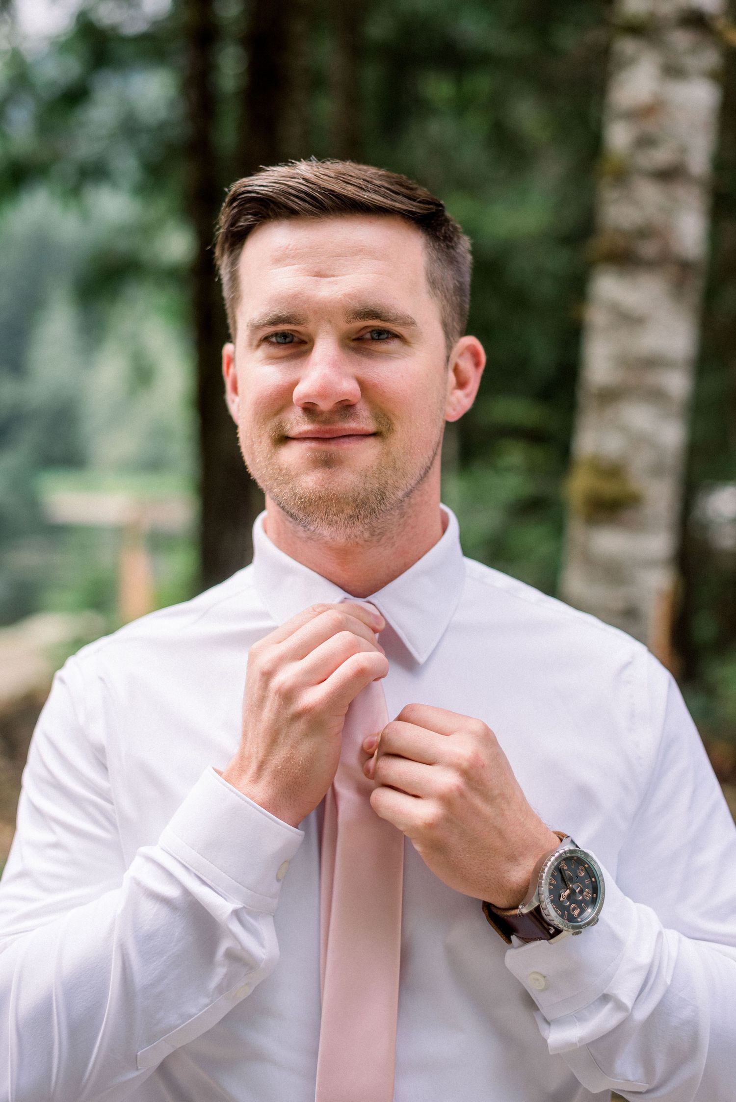 Groom in white shirt and pink tie smiling while getting ready outdoors.