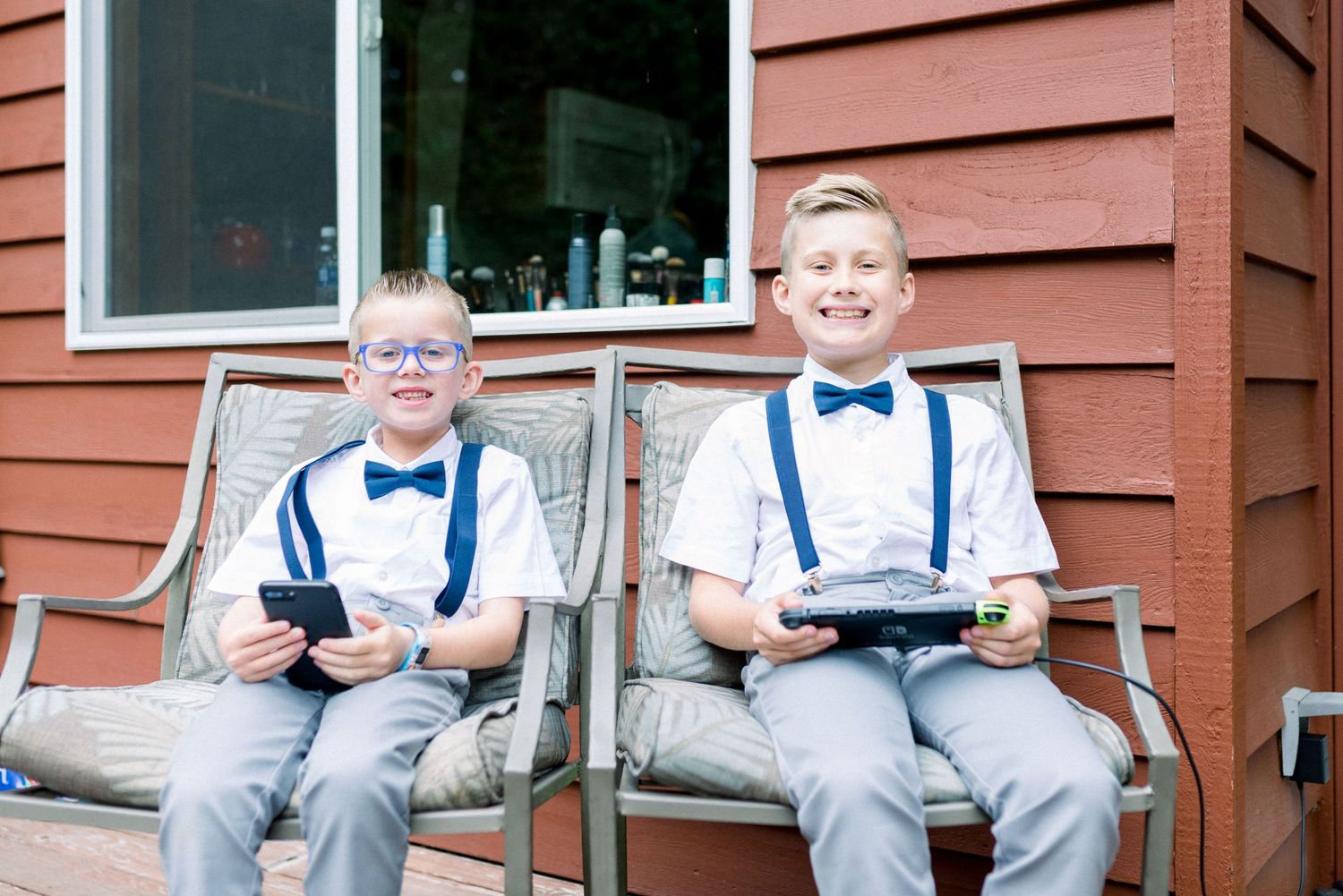 Two young ring bearers in matching bow ties and suspenders sitting on patio furniture.