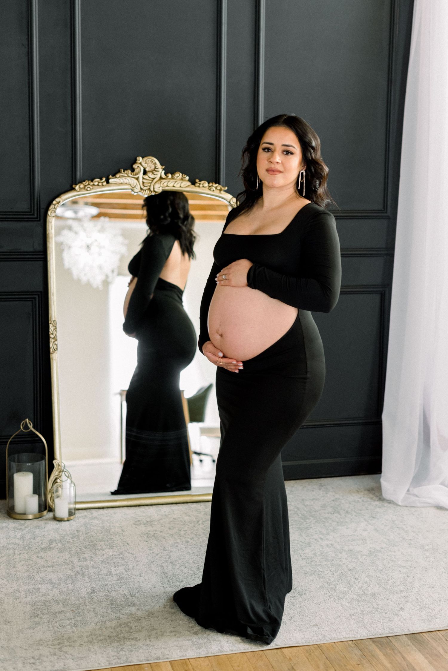Elegant maternity portrait featuring a black form-fitting dress and ornate gold mirror against dark paneled walls.