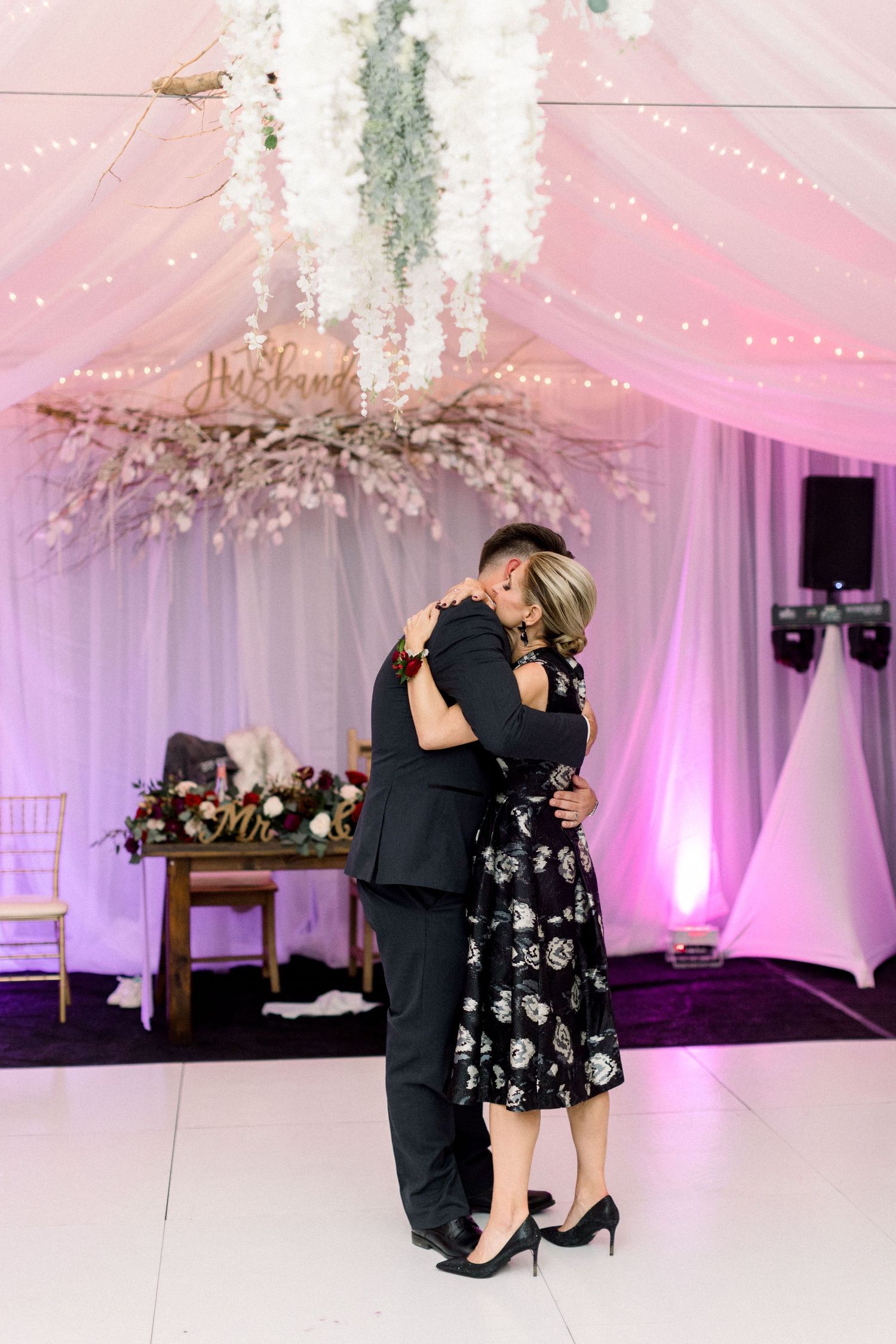 Couple shares intimate dance moment under hanging floral arrangements with purple lighting.