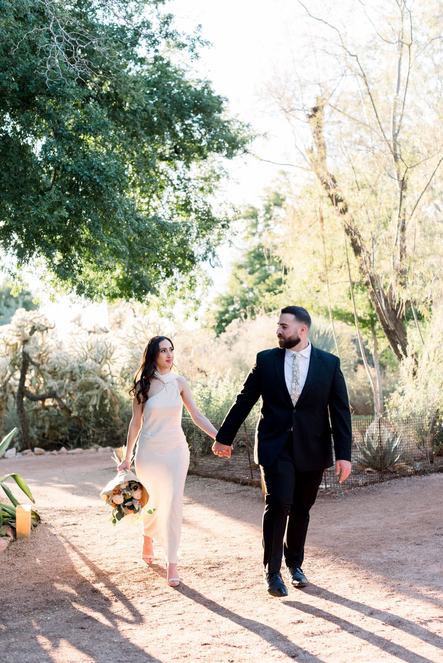 A couple walks hand in hand on a sunlit dirt path, with the bride holding a sunflower bouquet.