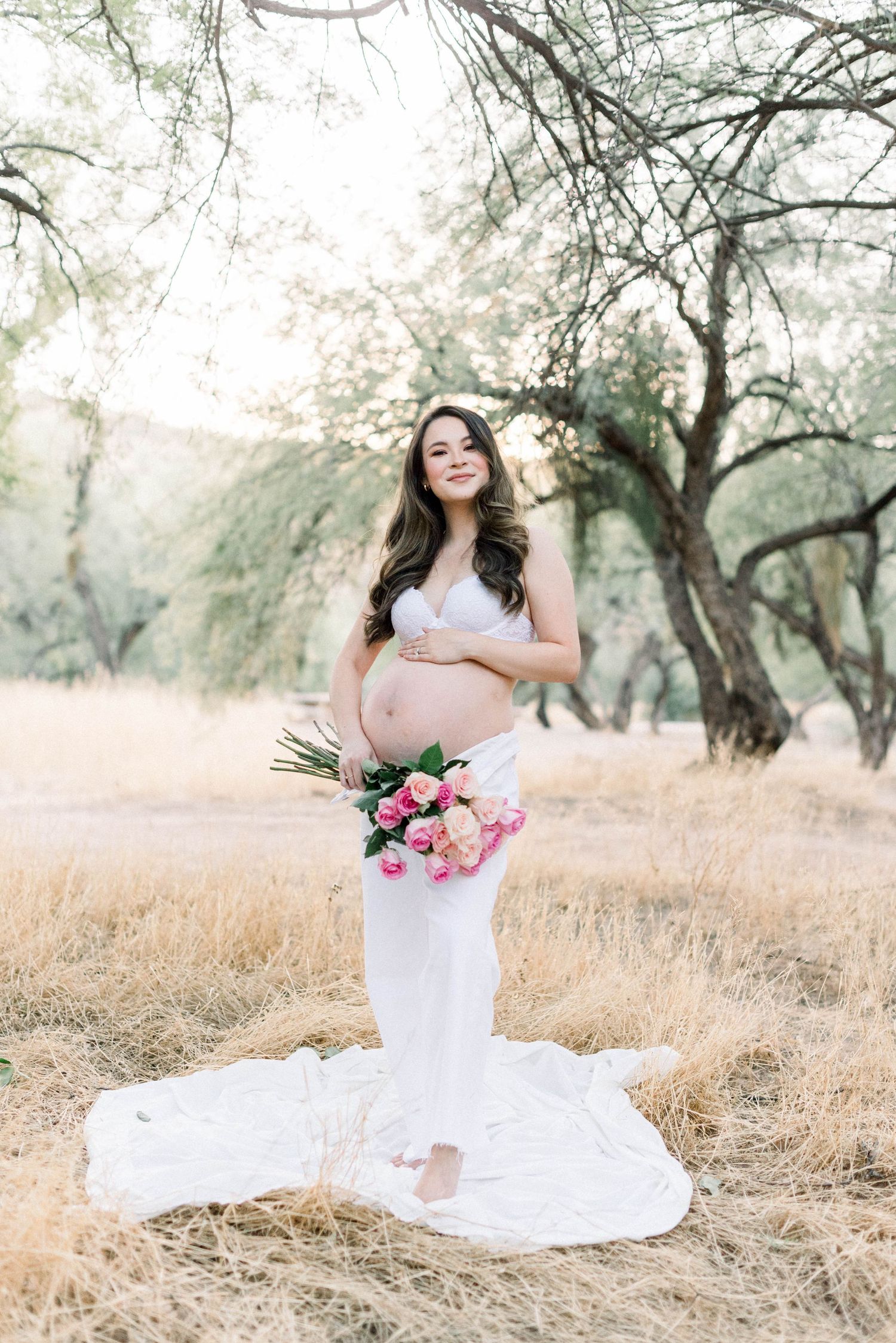 Maternity photo of woman in flowing white dress holding pink roses in dreamy outdoor setting with trees.