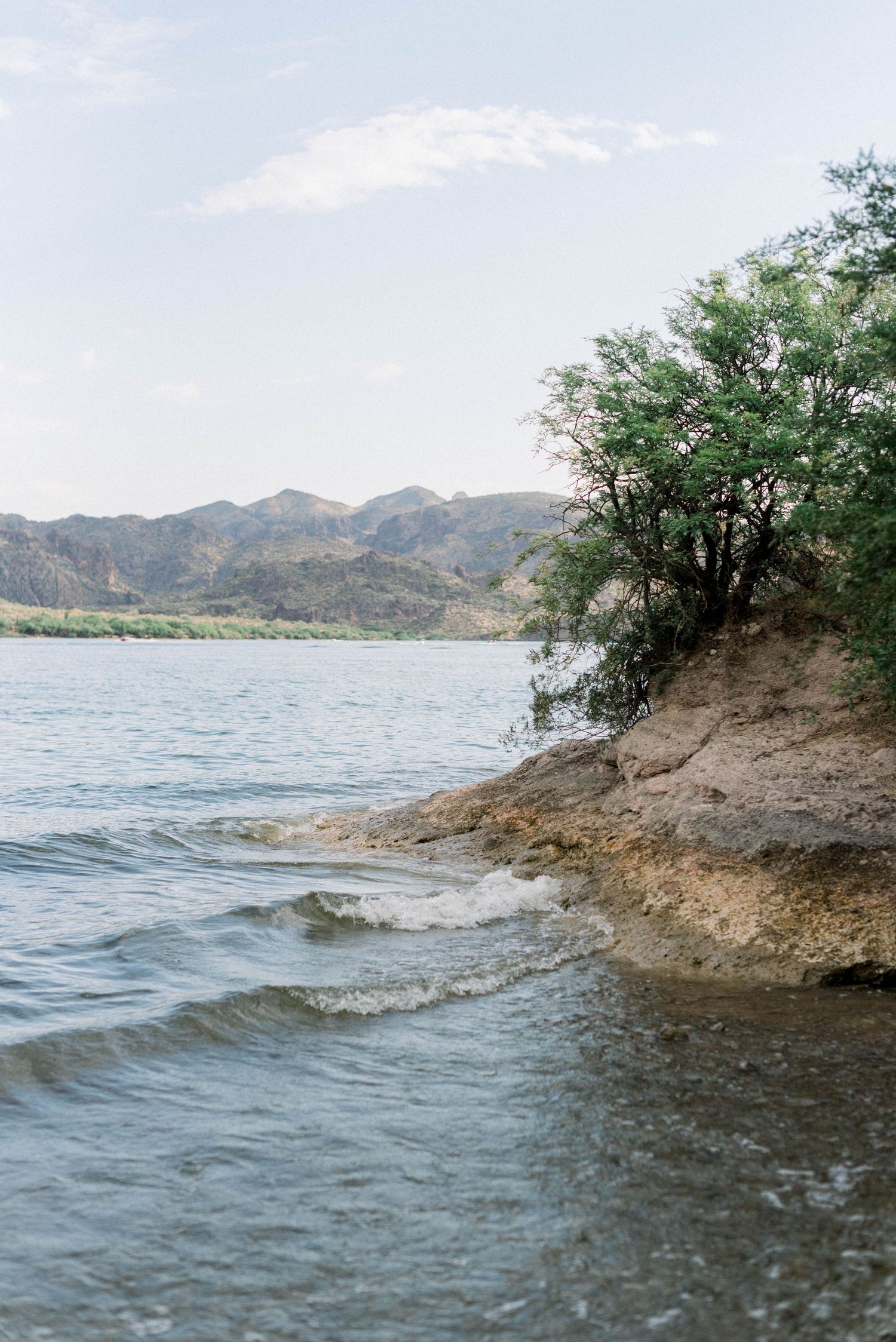 Scenic lake shoreline with rocky cliffs and trees against mountain backdrop during daytime.