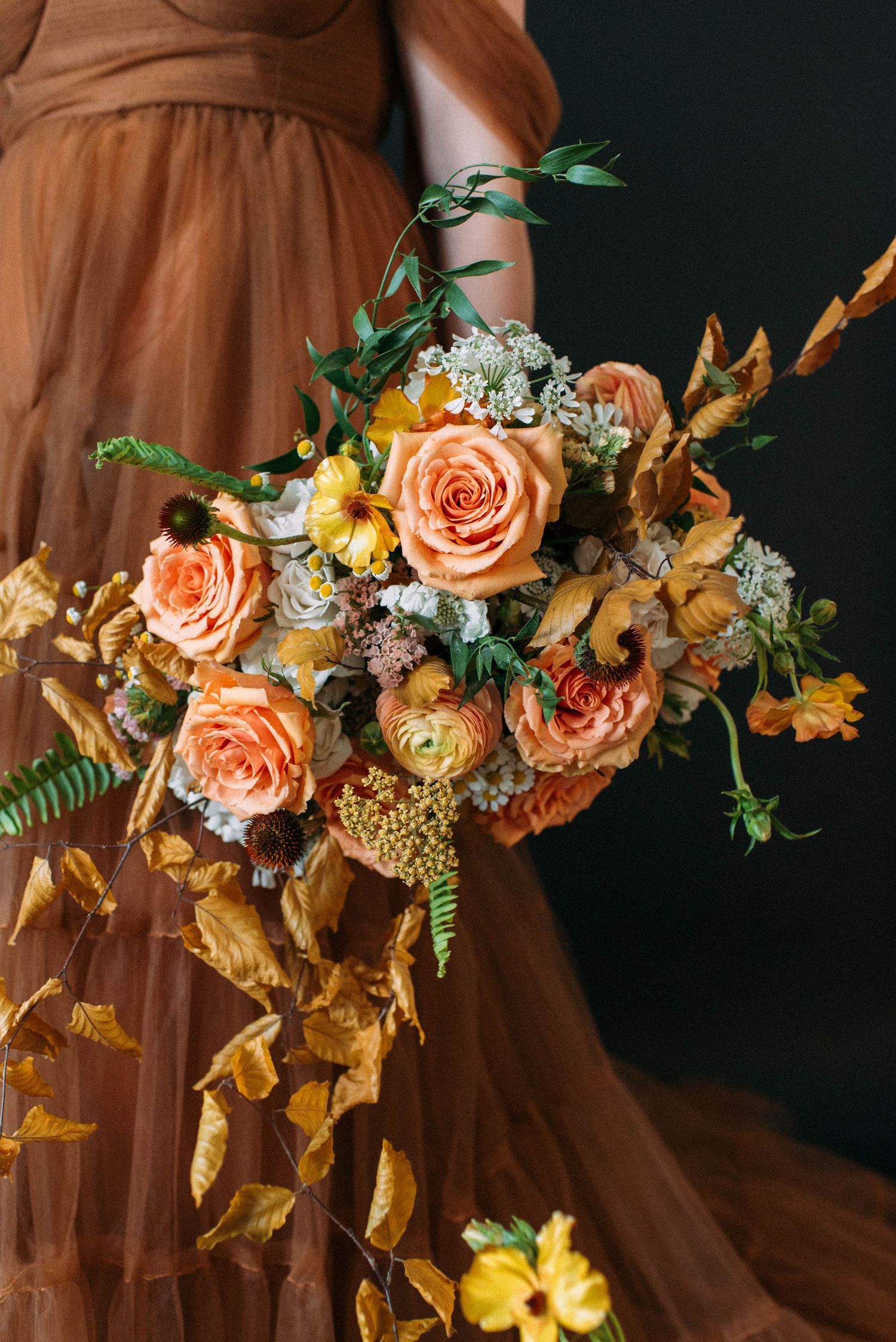 Autumn-inspired floral arrangement with peach roses, golden leaves, and delicate ferns against a dark background.