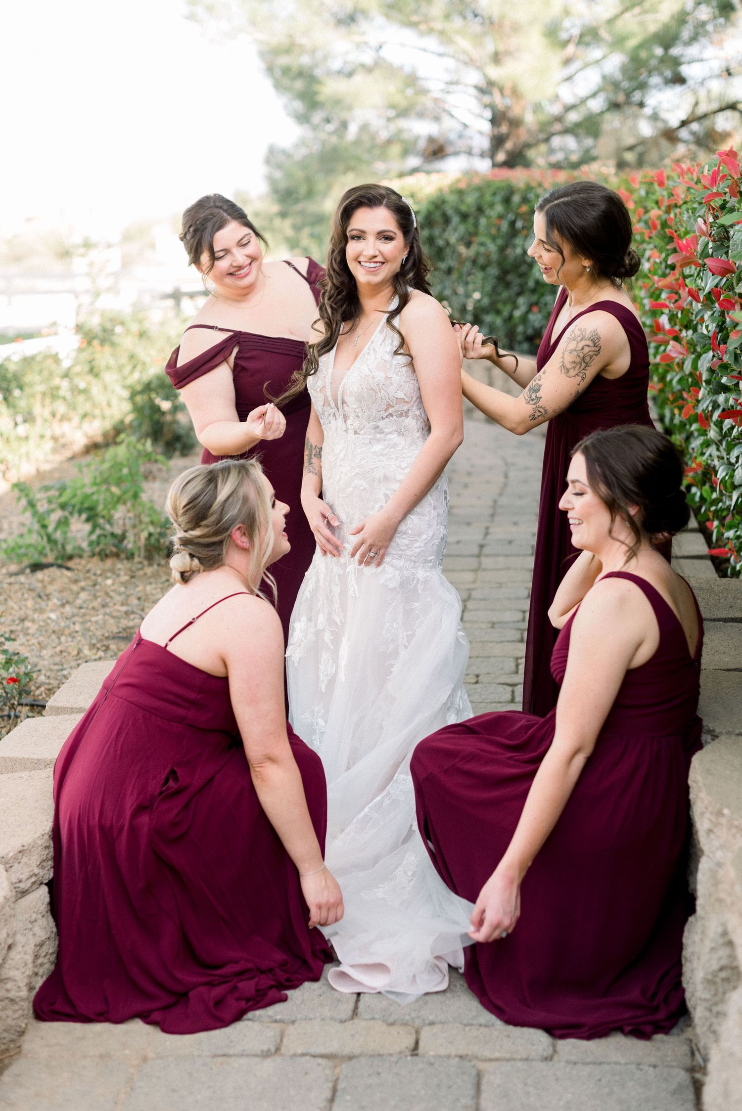 Bridesmaids in burgundy dresses help arrange the train of a white lace wedding gown on stone steps.