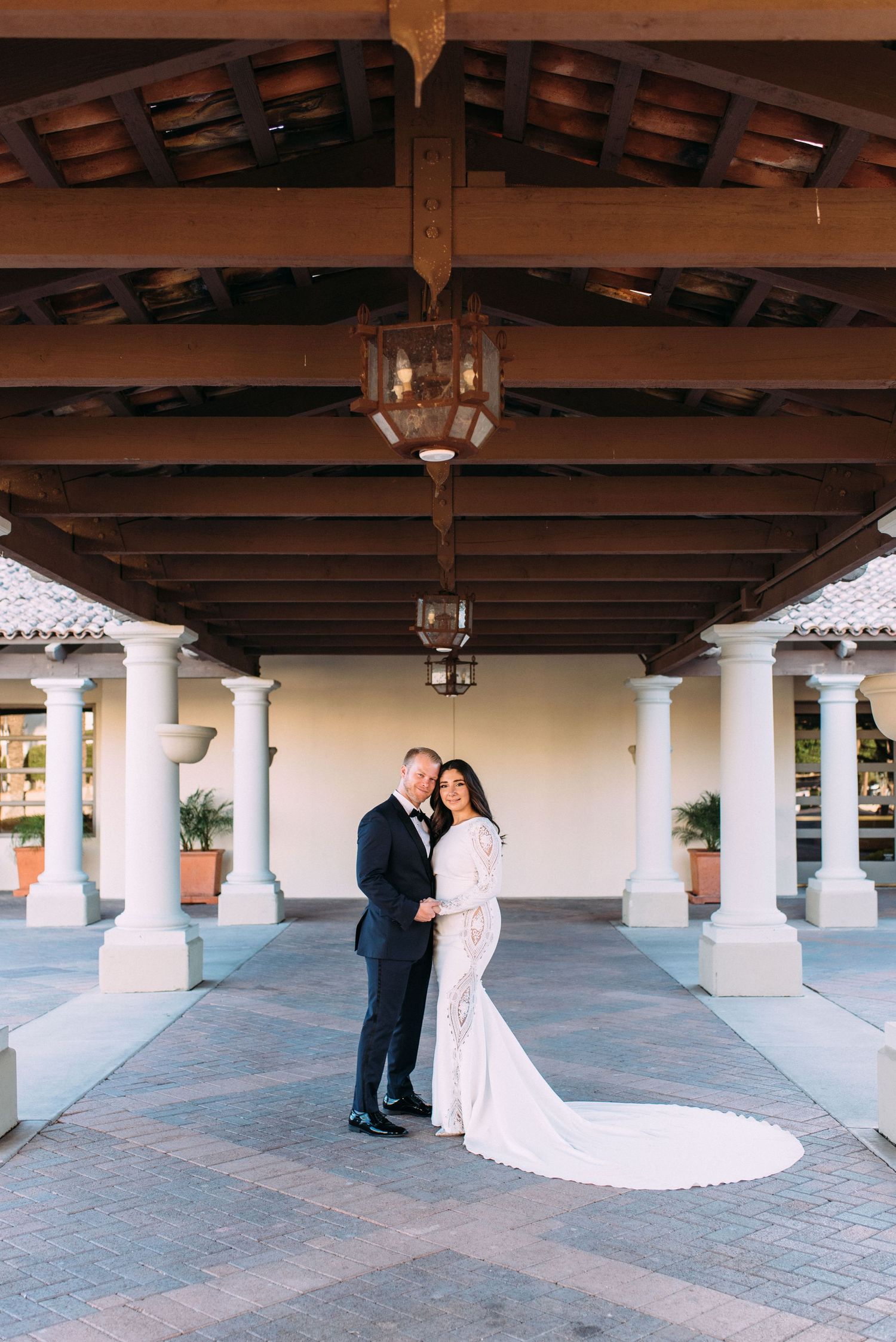 A couple embraces under an elegant covered portico with white columns and exposed wooden beams.