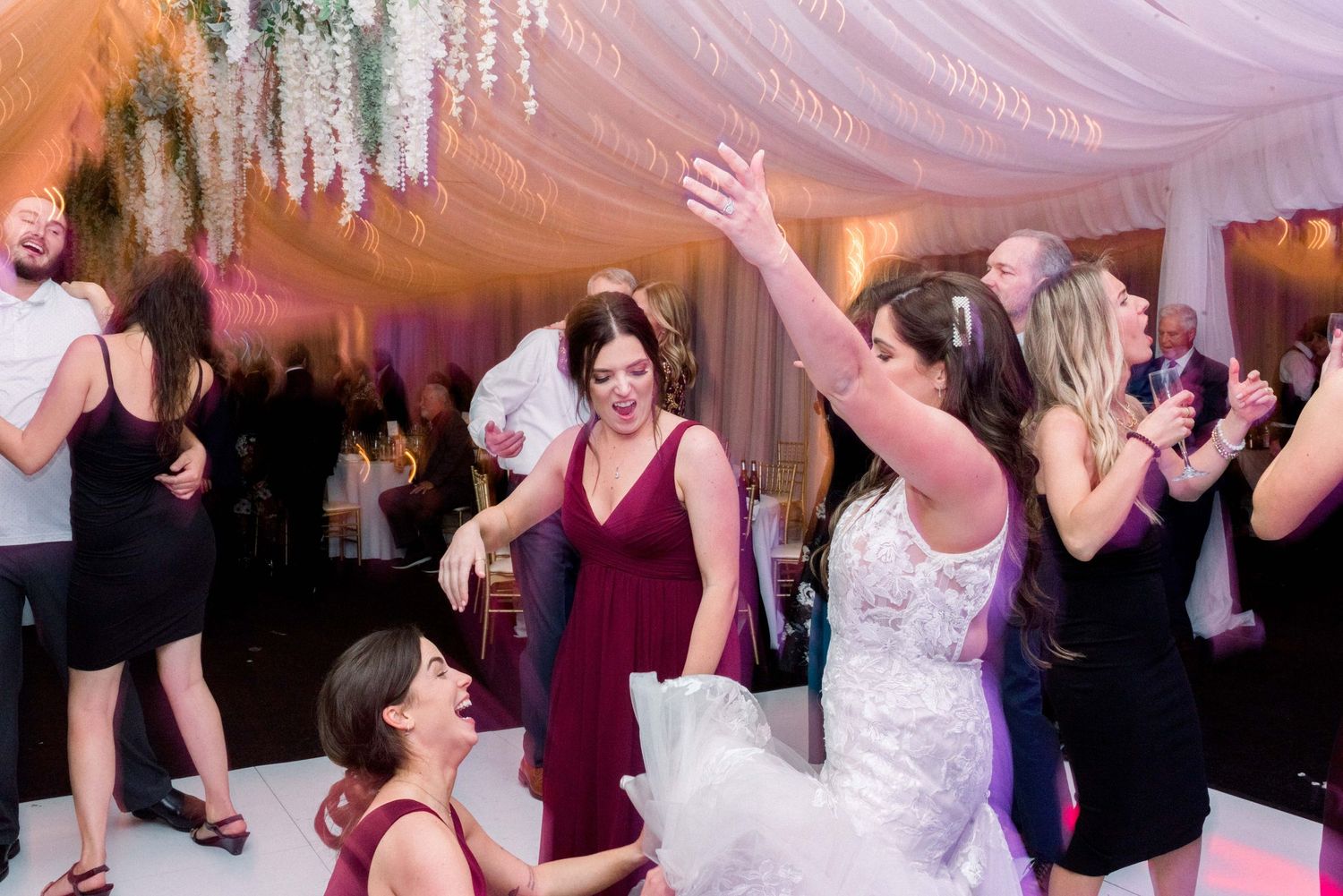 Wedding party celebrating on dance floor with raised arms under elegant draped ceiling.