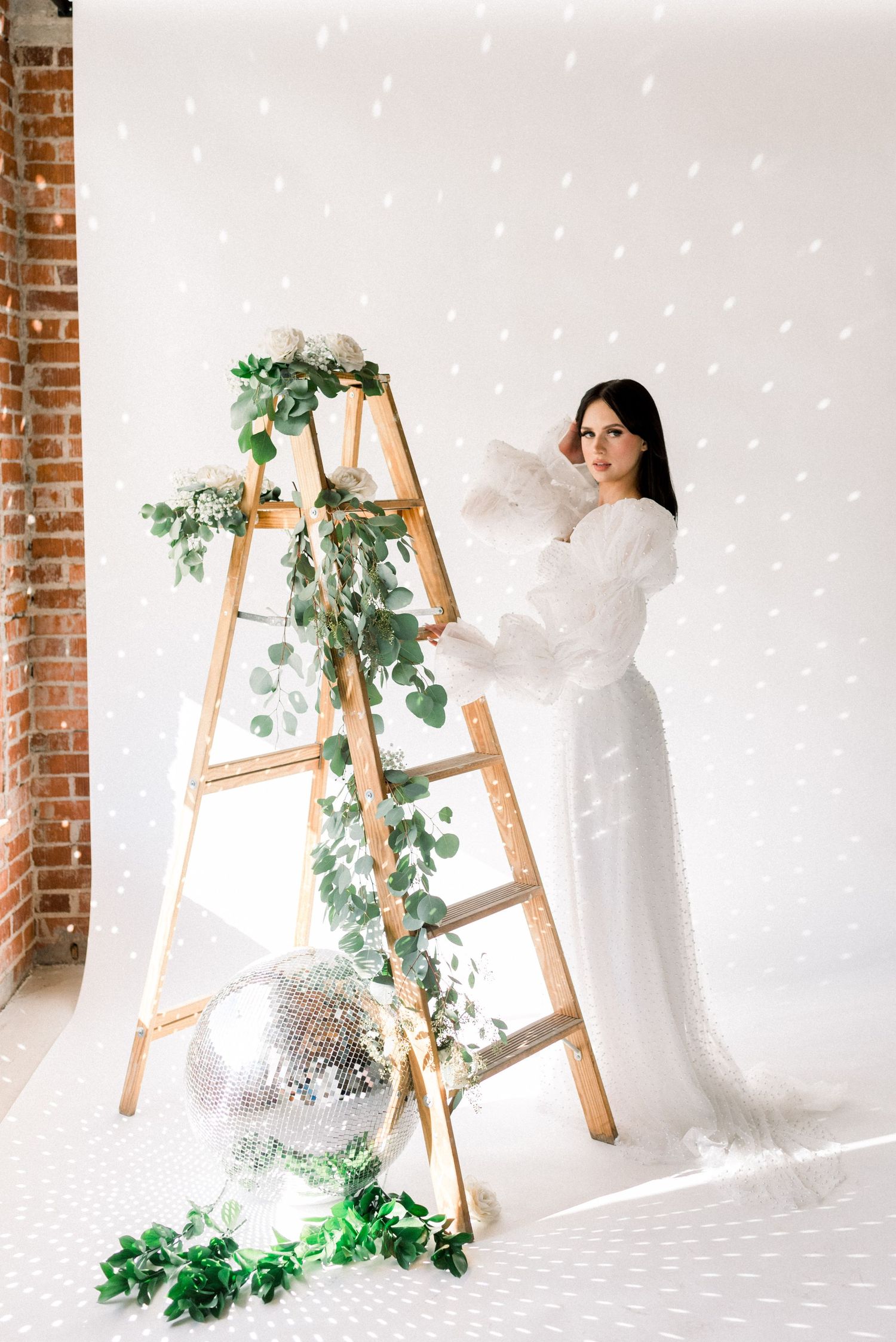 Fashion model in flowing white dress poses next to wooden ladder decorated with holiday greenery.