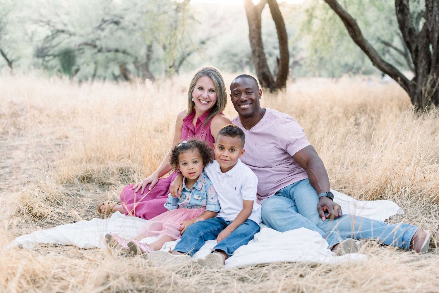 A family sits together on a white blanket in a golden grass field during sunset.