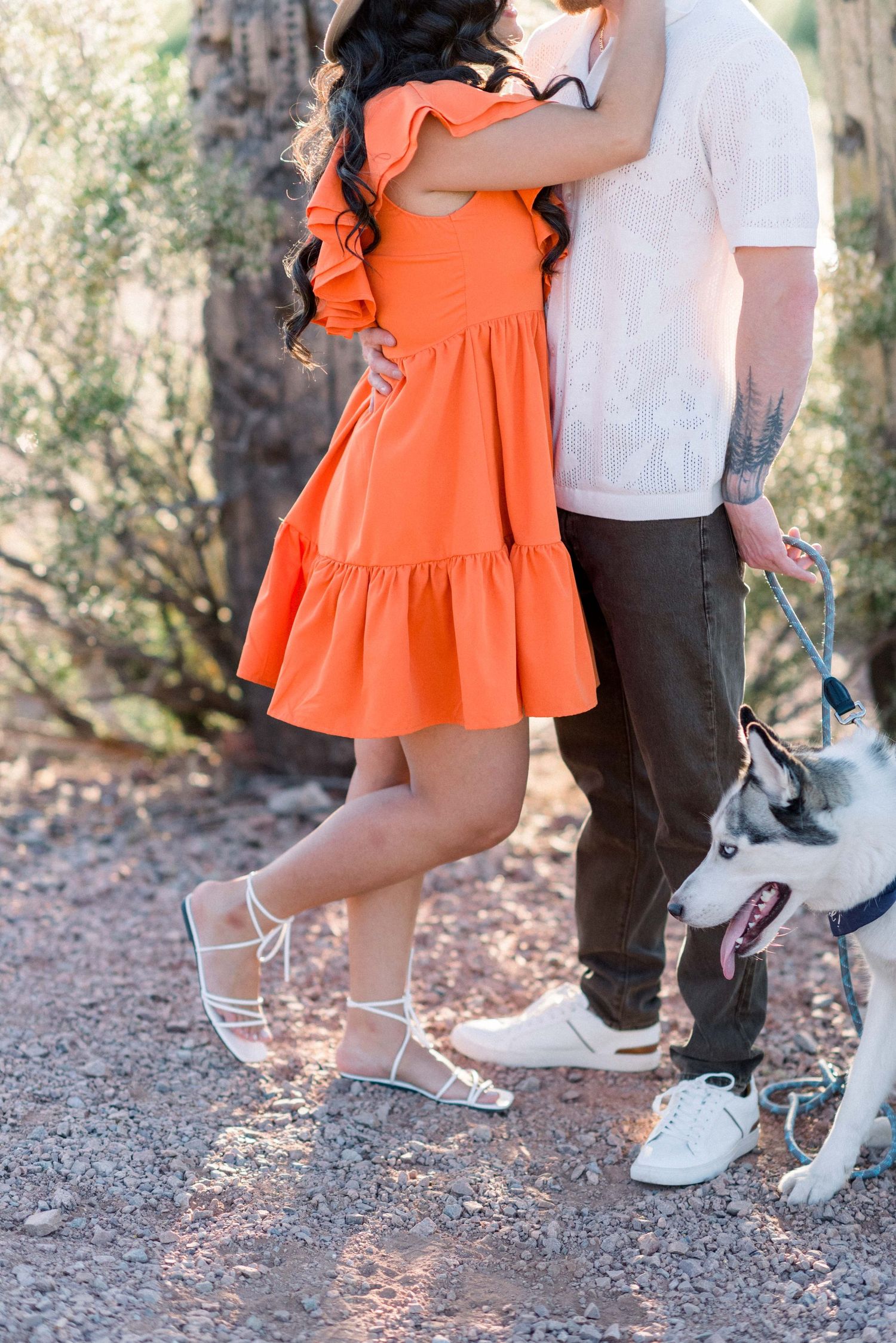 A couple stands close together on a trail with their dog, focusing on their feet and orange dress details.