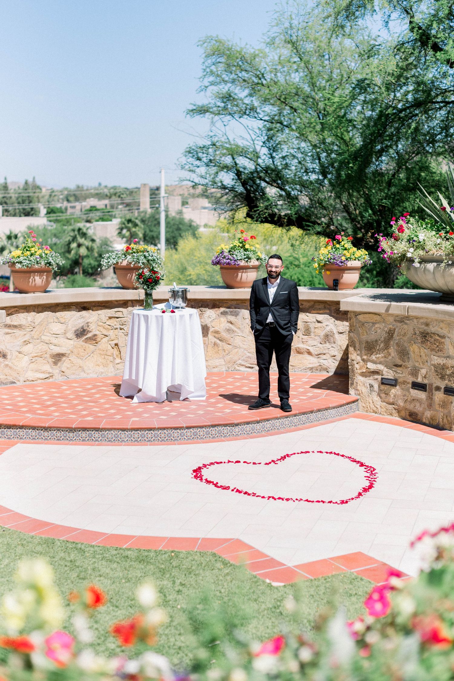 Groom standing on patio with heart shape made of rose petals during proposal setup.