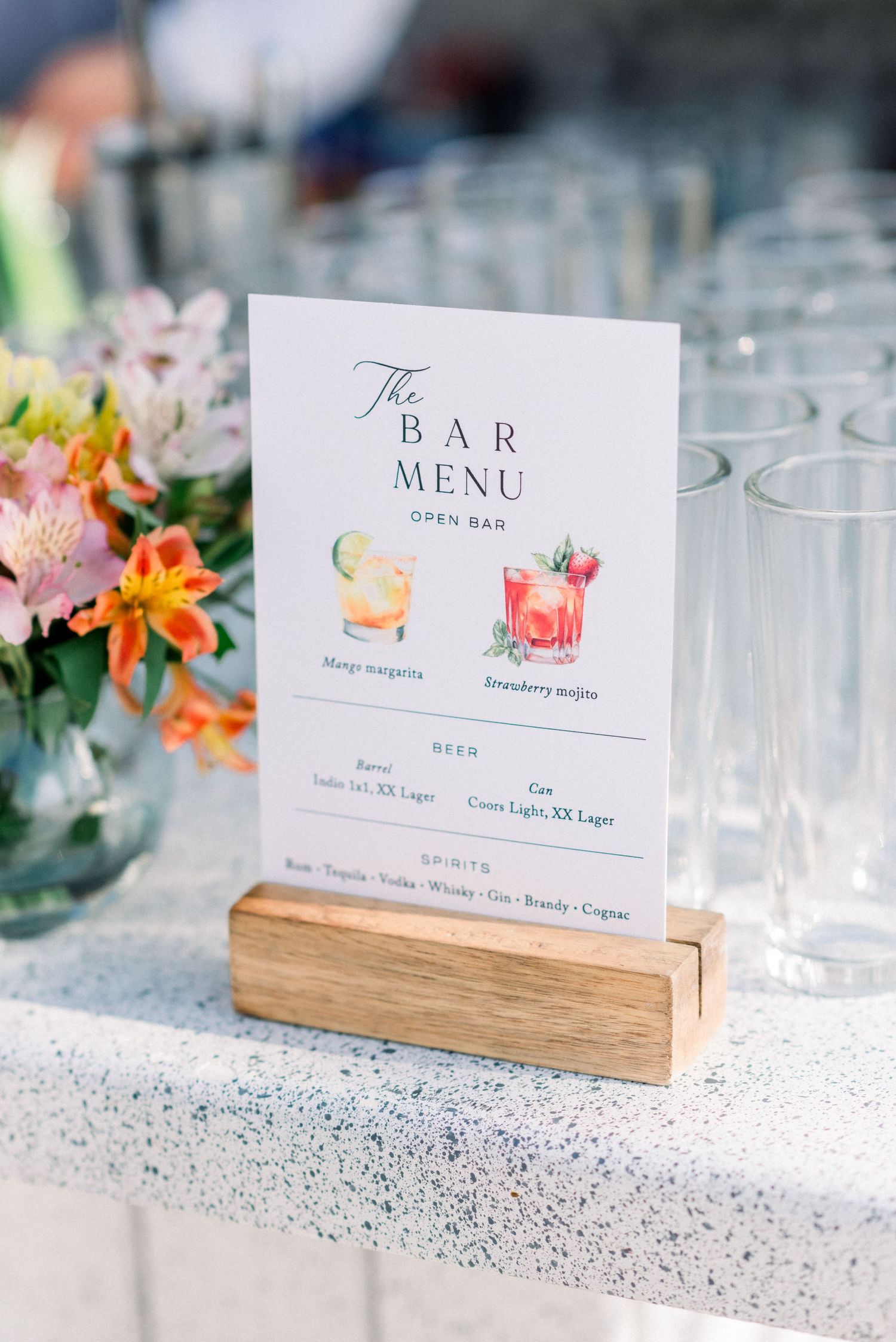 Bar menu display with cocktail illustrations in wooden stand next to floral centerpiece.