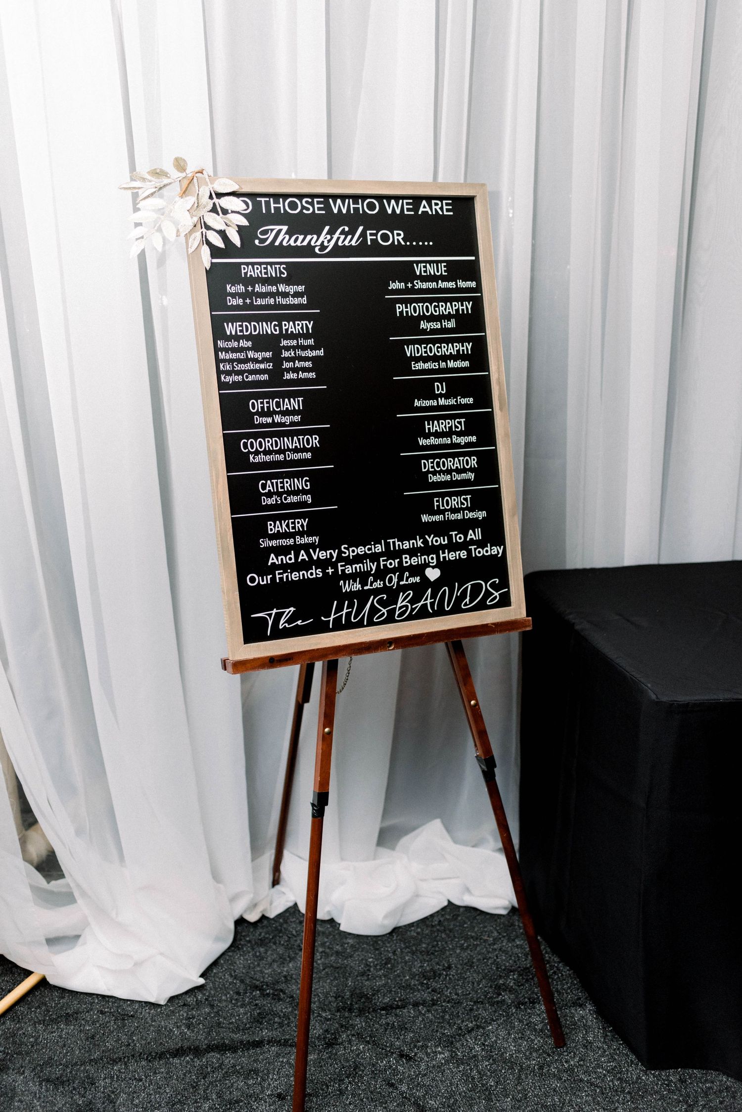 A chalkboard wedding sign on an easel displays 'Thankful For' message against white drapes.
