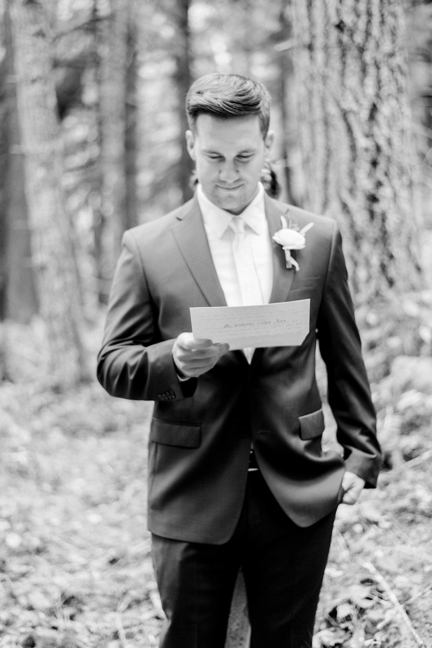 A groom in a dark suit reads a letter in a wooded setting, wearing a boutonniere.