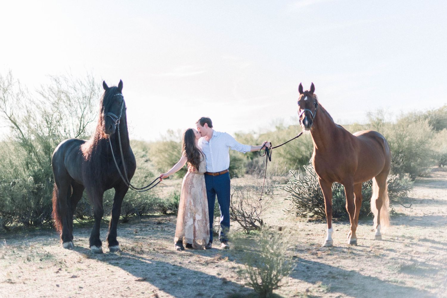 Couple posing for engagement photos with two horses in a desert landscape.