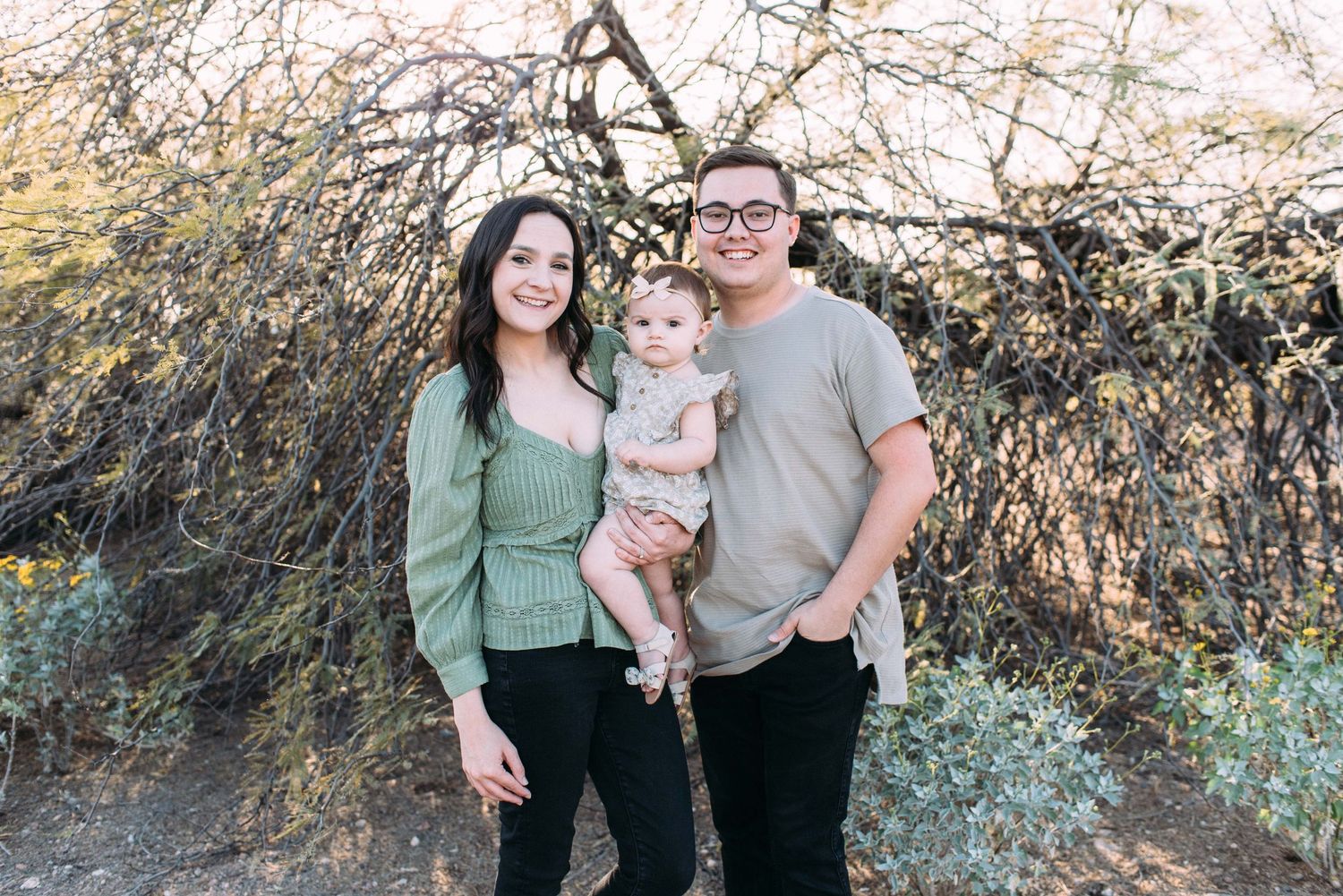 Family of three standing together on desert path with brush and branches.