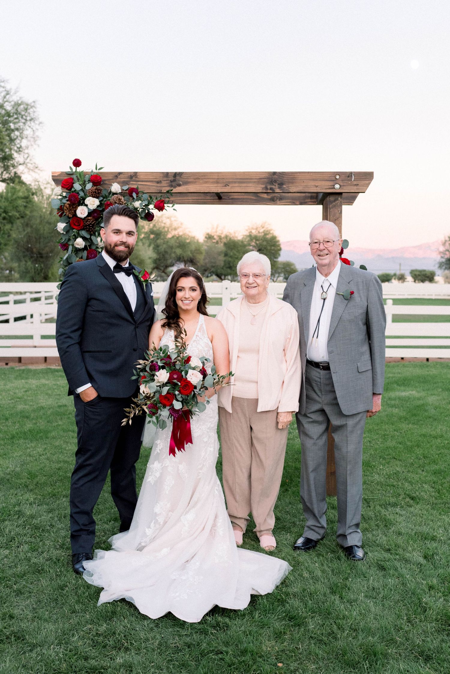 Outdoor wedding portrait with newlyweds and family members under a wooden pergola decorated with red roses.