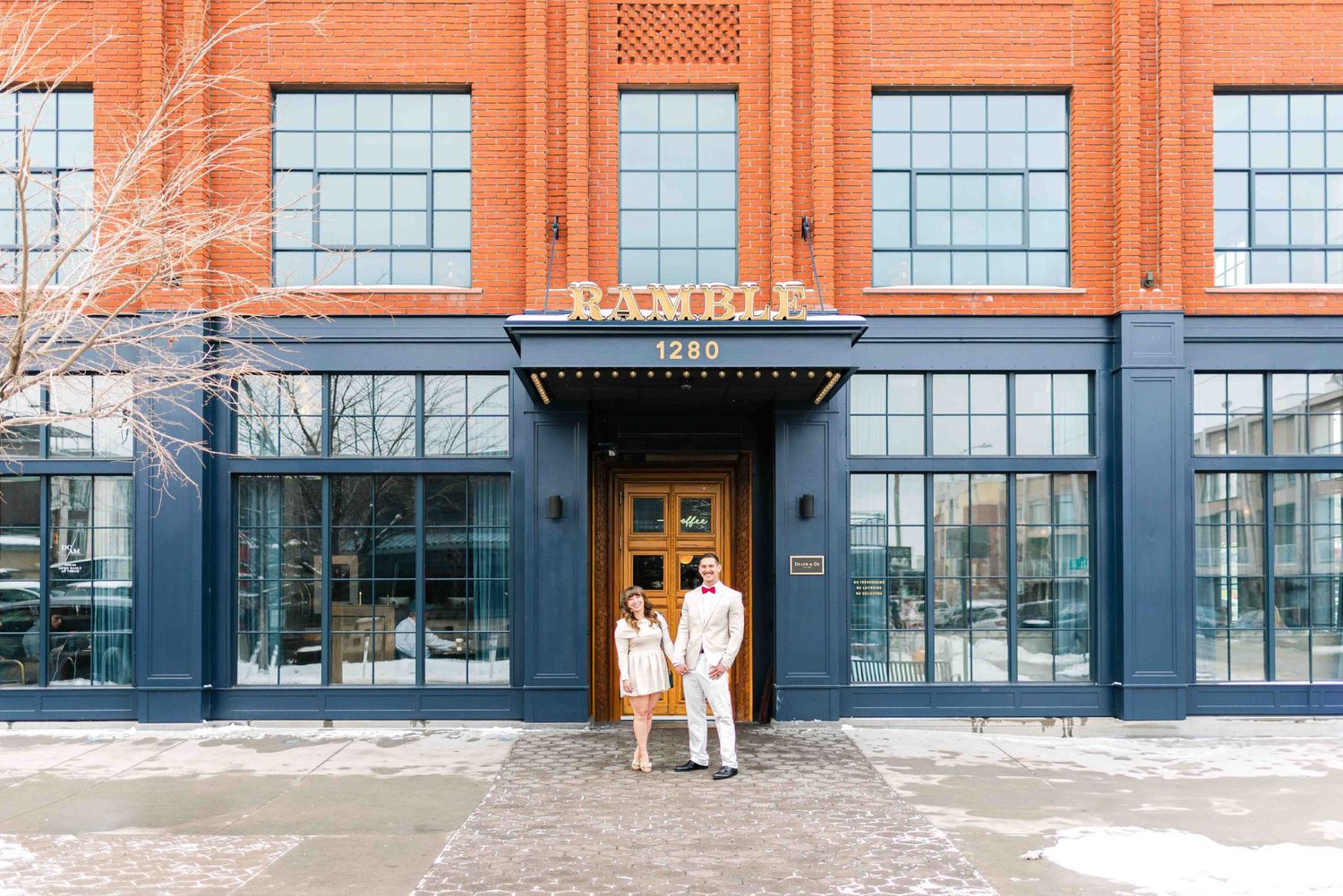 A couple poses in white outfits outside a modern industrial-style building entrance.