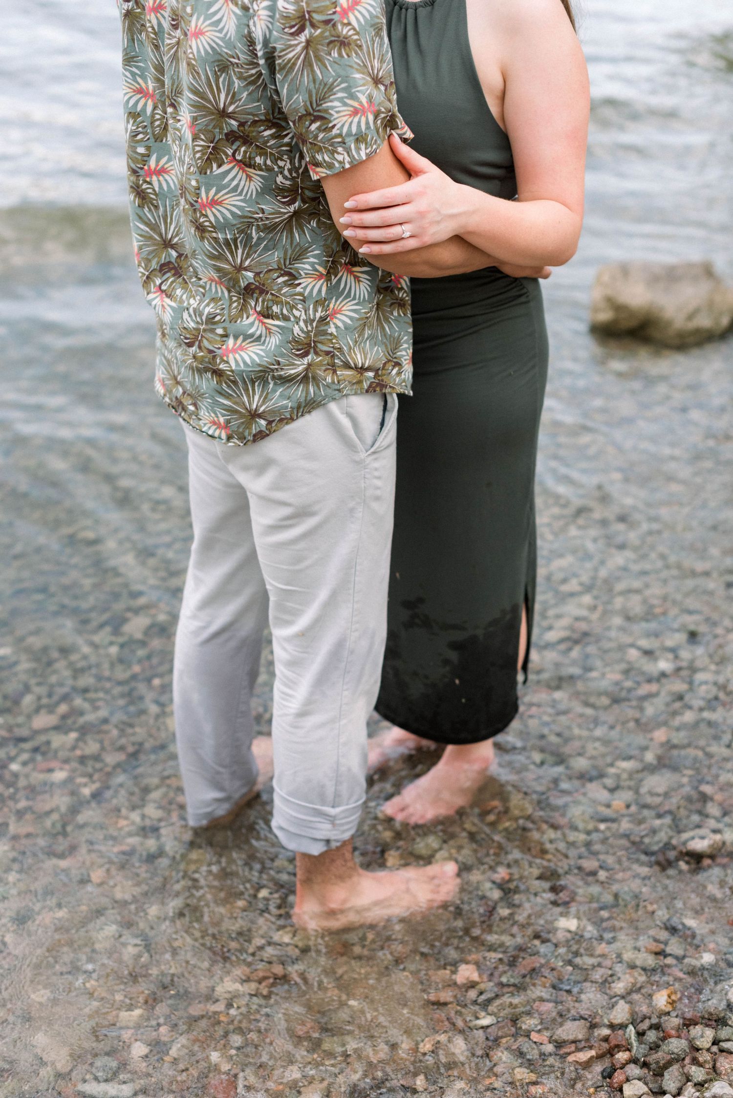 Close up of a couple's legs standing barefoot on a rocky lakeshore wearing floral shirt and dark dress.