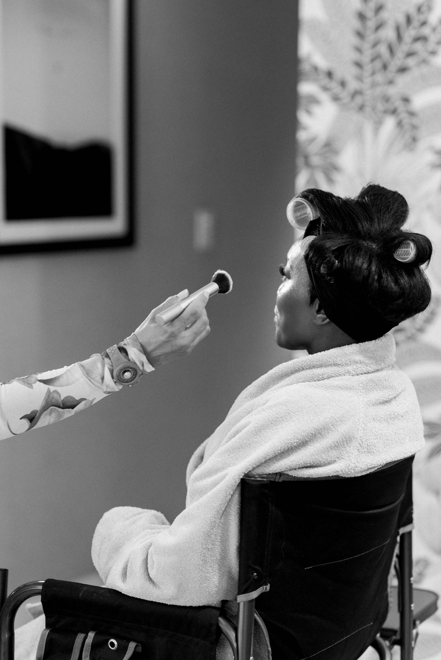A person in a white robe sits in a chair while someone with tattooed arms applies makeup in black and white photography.
