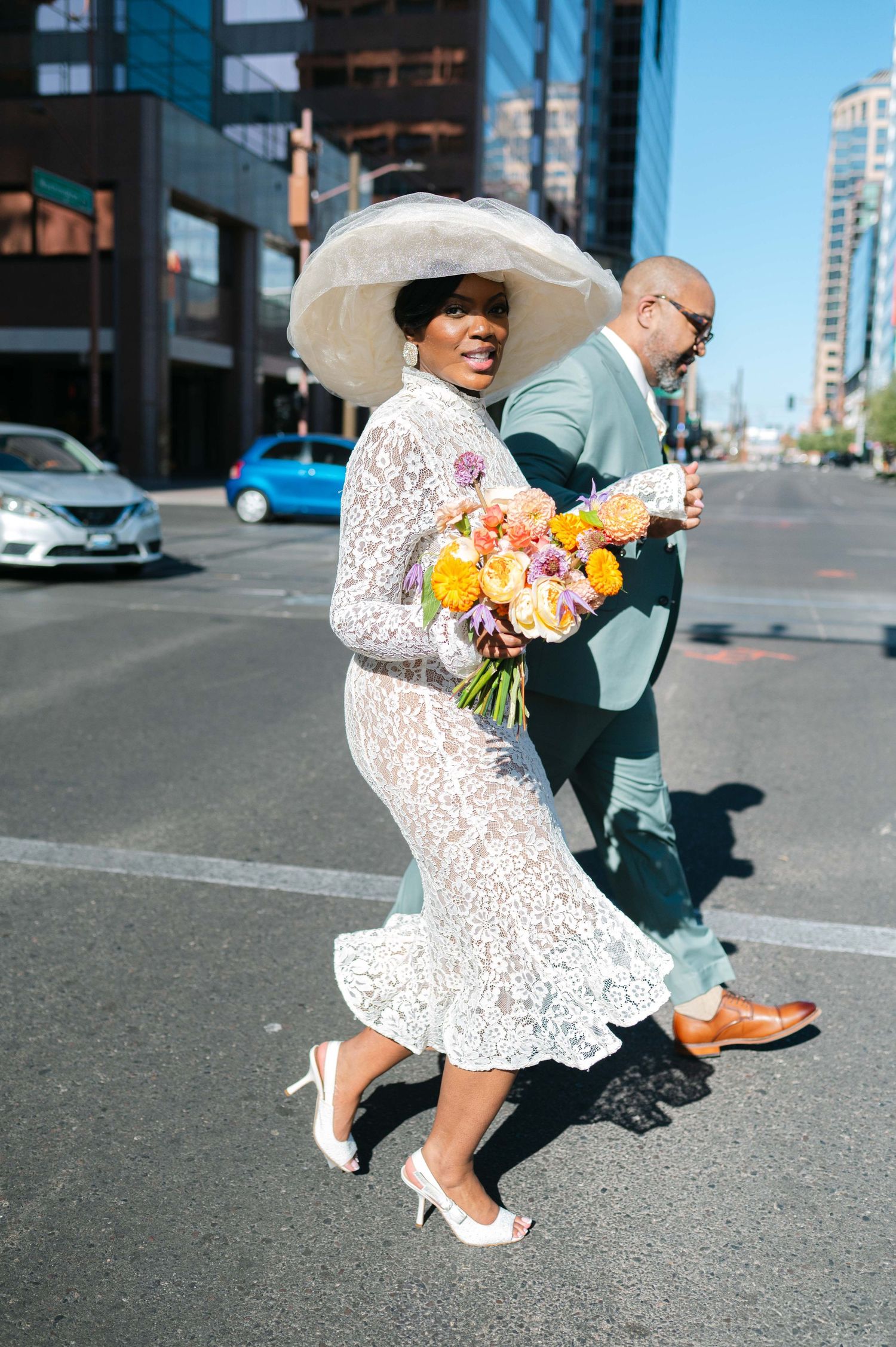 A couple in formal attire dances playfully on a city street with colorful flowers.
