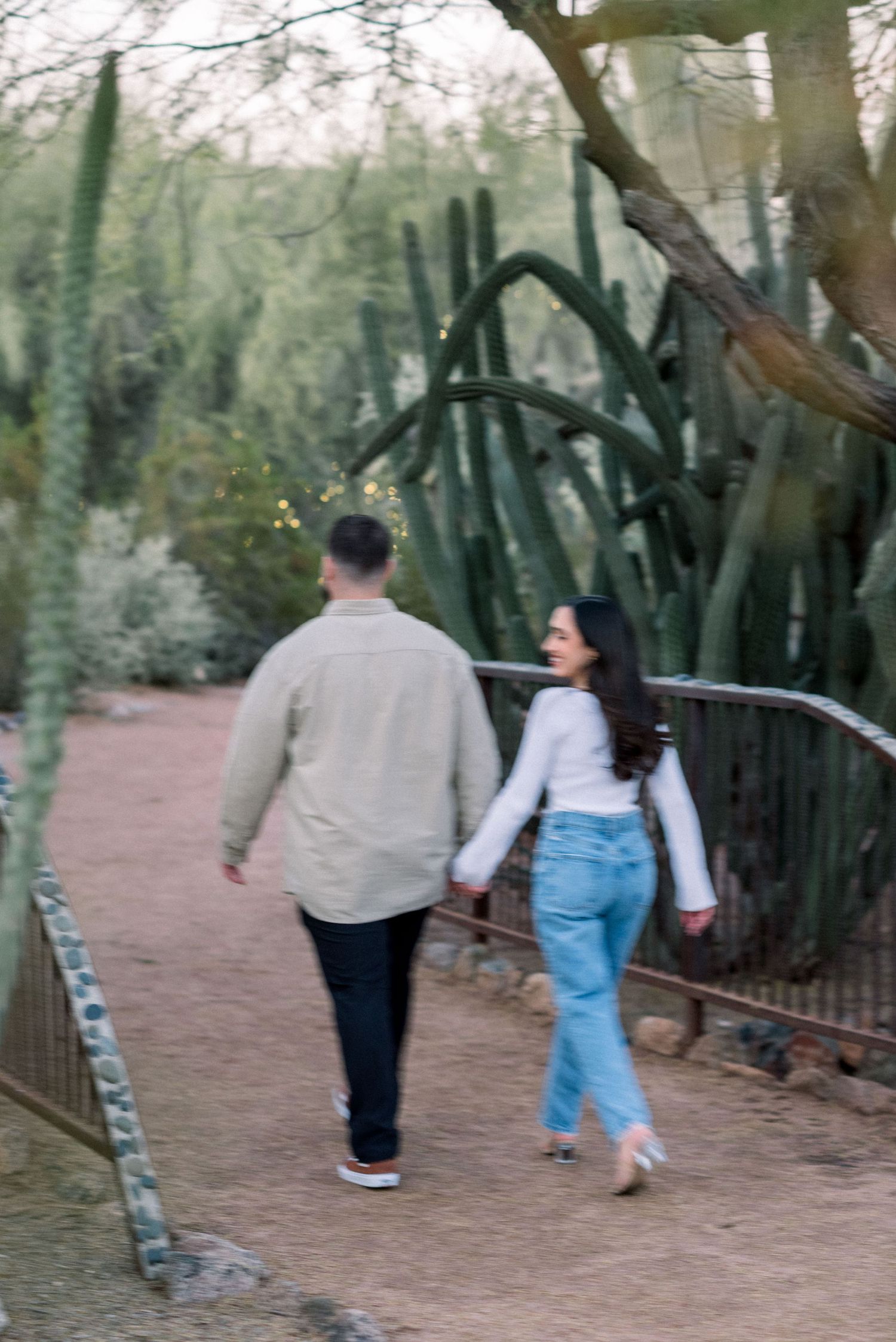 A couple walks together on a dirt path through a desert garden with tall cacti and trees.