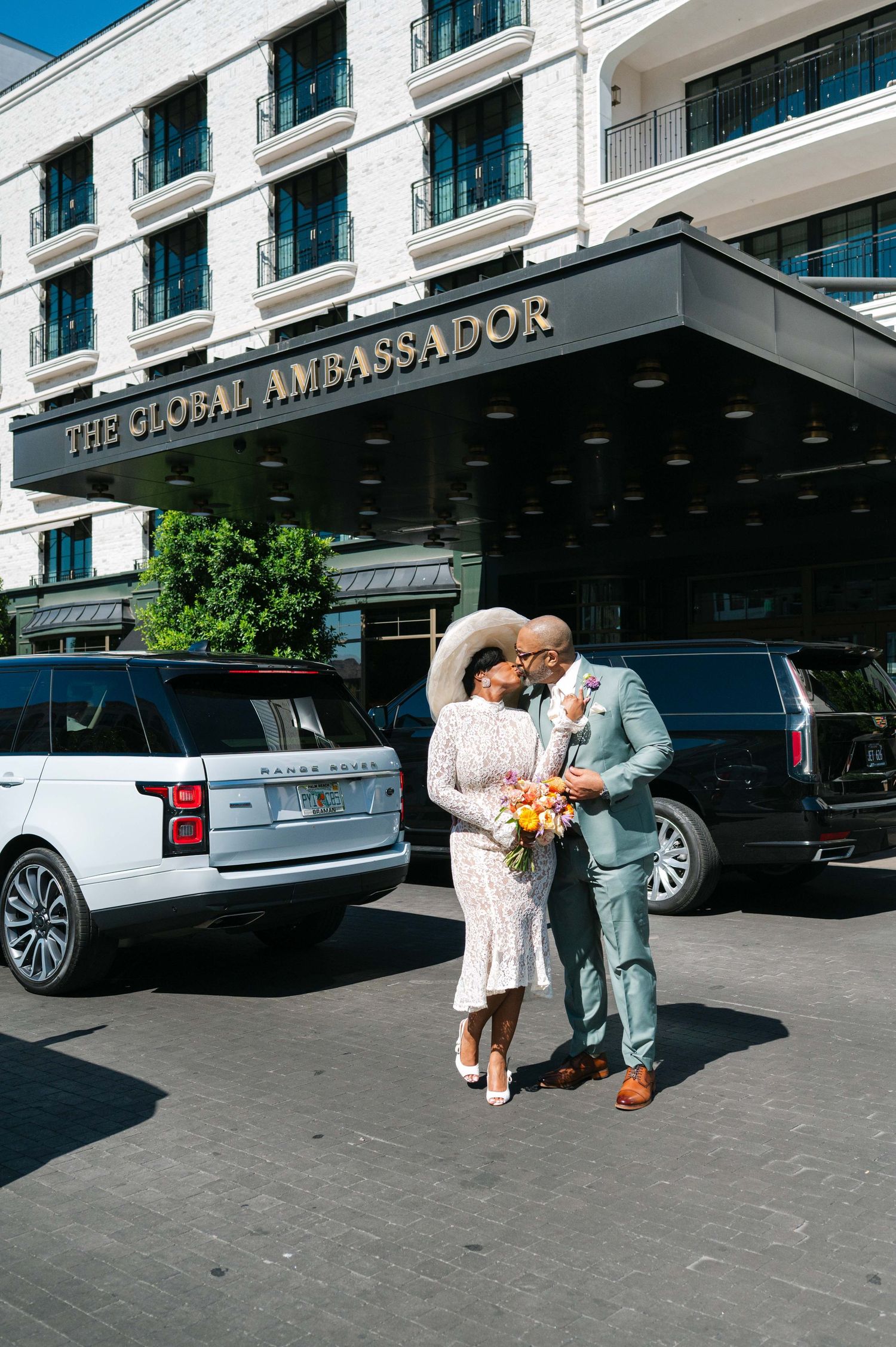 Wedding couple embracing outside The Grand Ambassador hotel entrance with luxury cars in background.