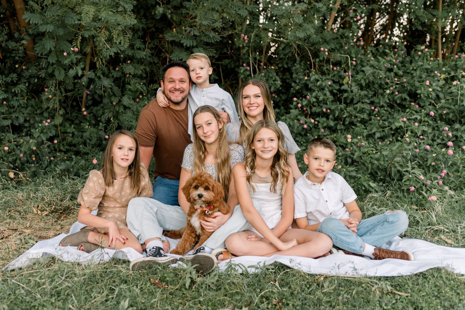 A family with a brown dog sits together on a white blanket in a wooded outdoor setting during a photo session.