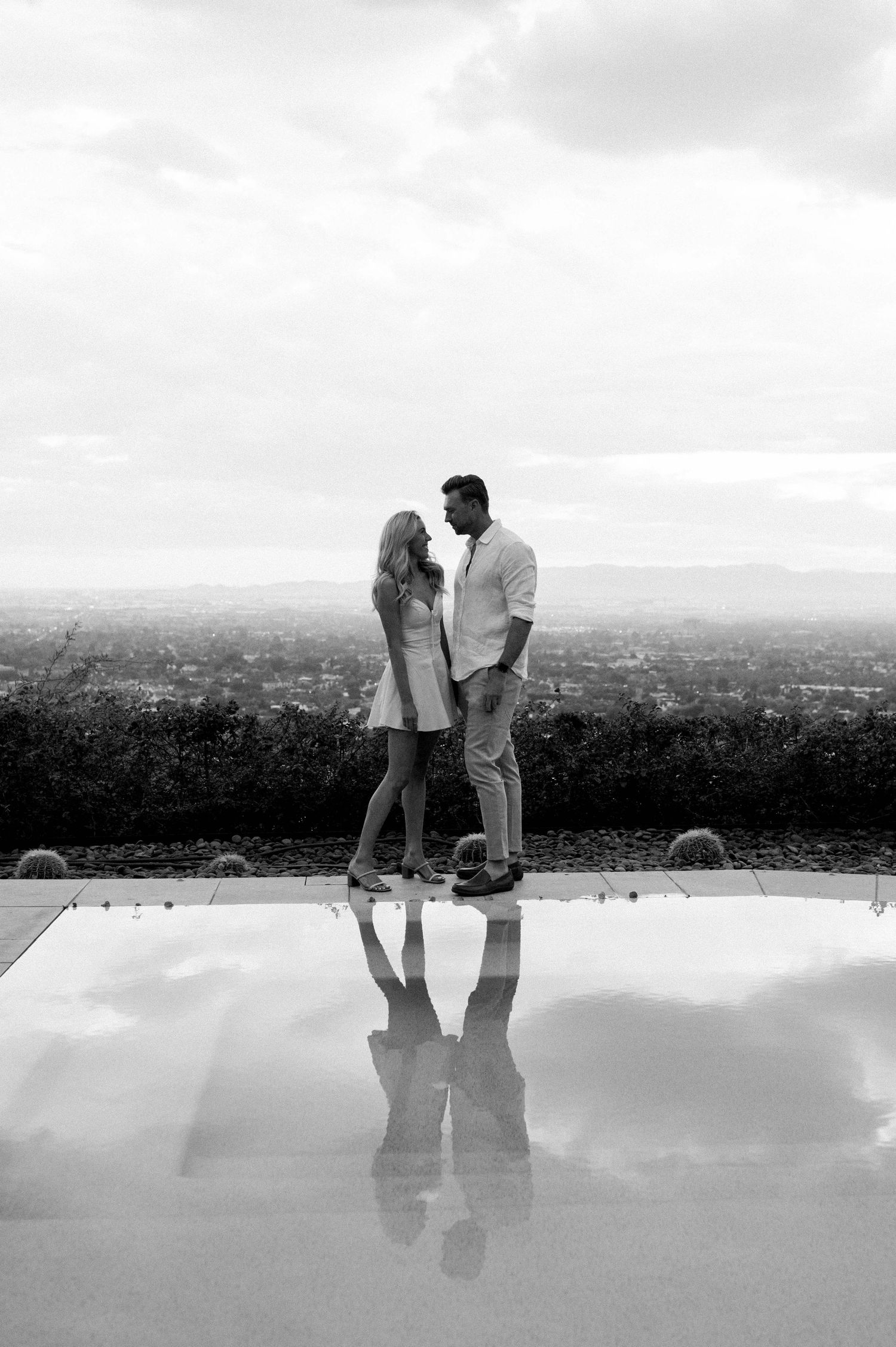 A black and white photo capturing reflections of a couple in a rooftop pool with city views.