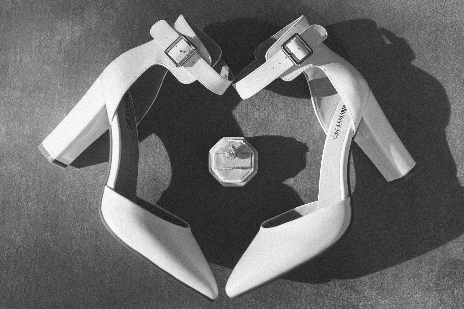 White bridal shoes arranged in heart shape with ring in black and white photography.