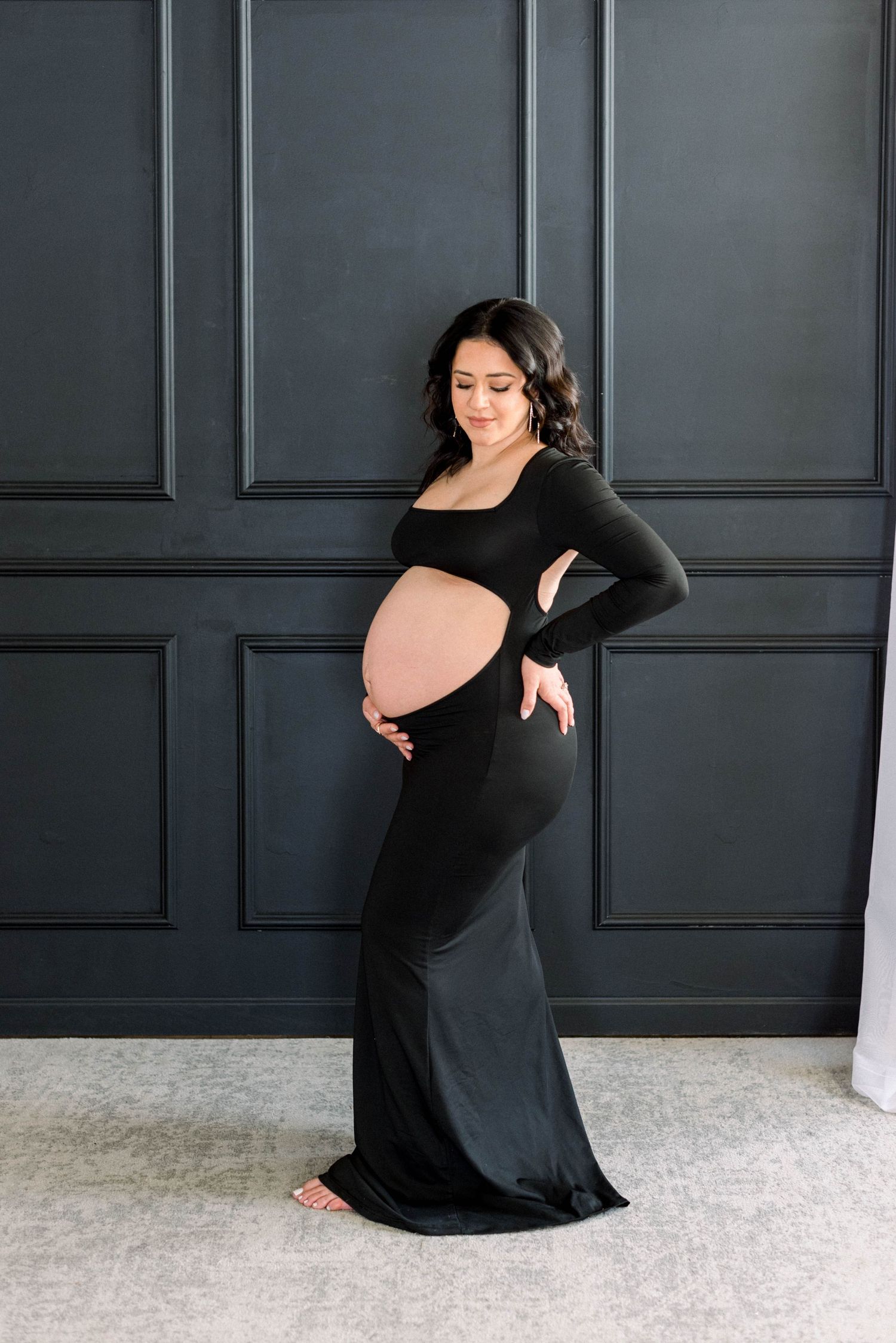 Elegant maternity pose in black form-fitting dress against dark paneled background.