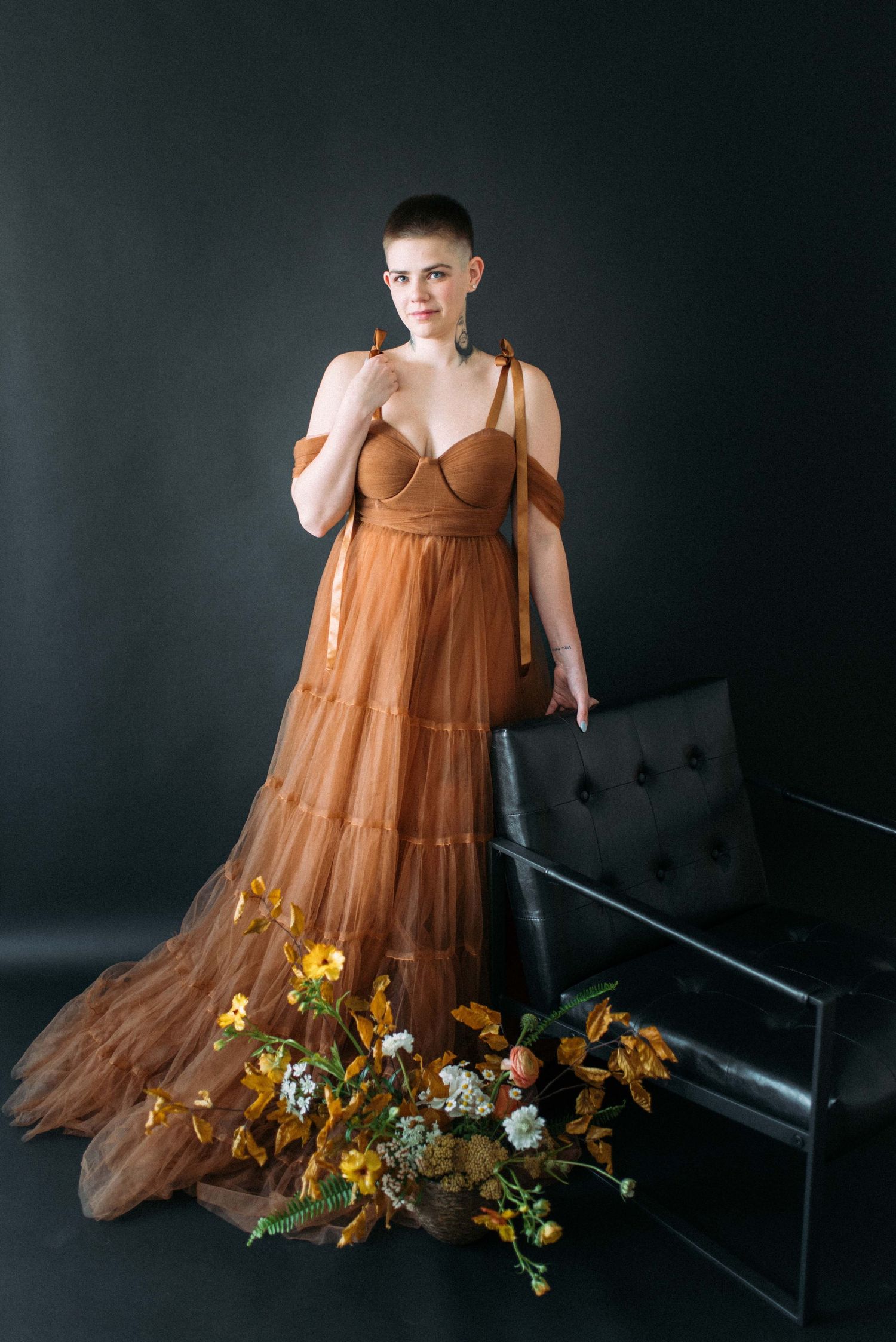 A formal portrait with yellow flowers beside a black leather bench in a brown flowy dress.