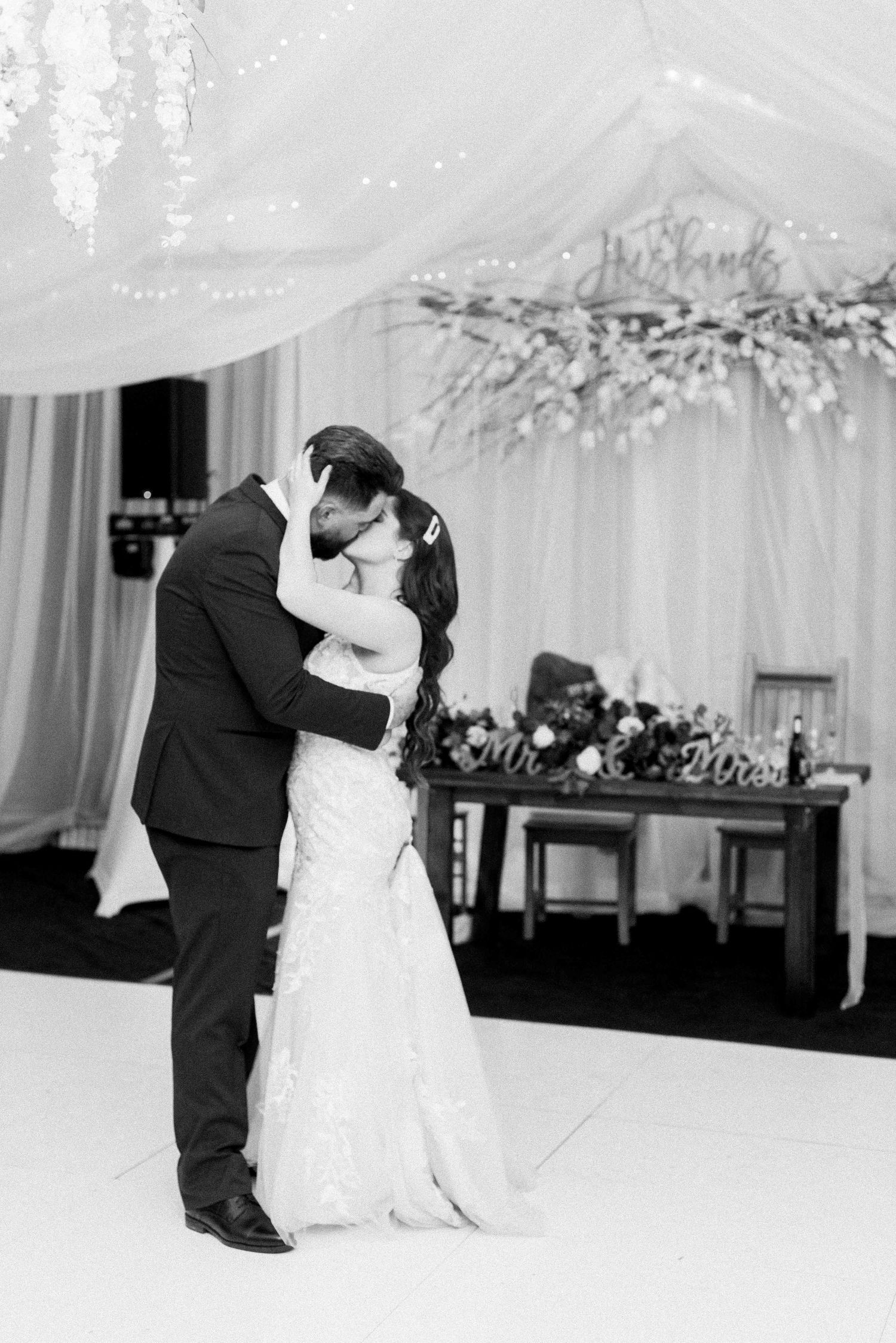 A black and white photograph of a romantic first dance at a wedding reception under draped fabric and chandeliers.