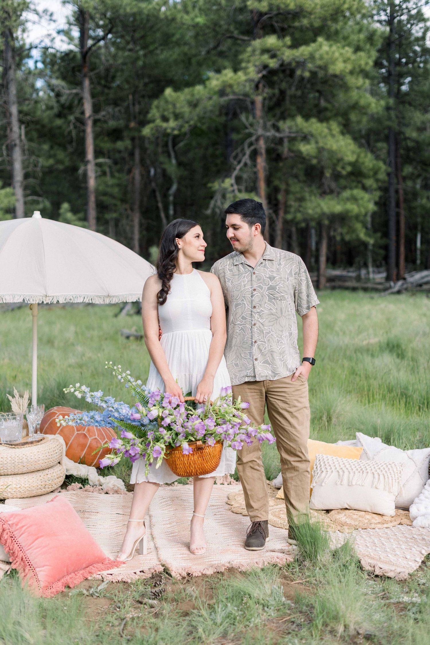 Couple enjoying a romantic picnic setup with flowers and cushions in a forest clearing.